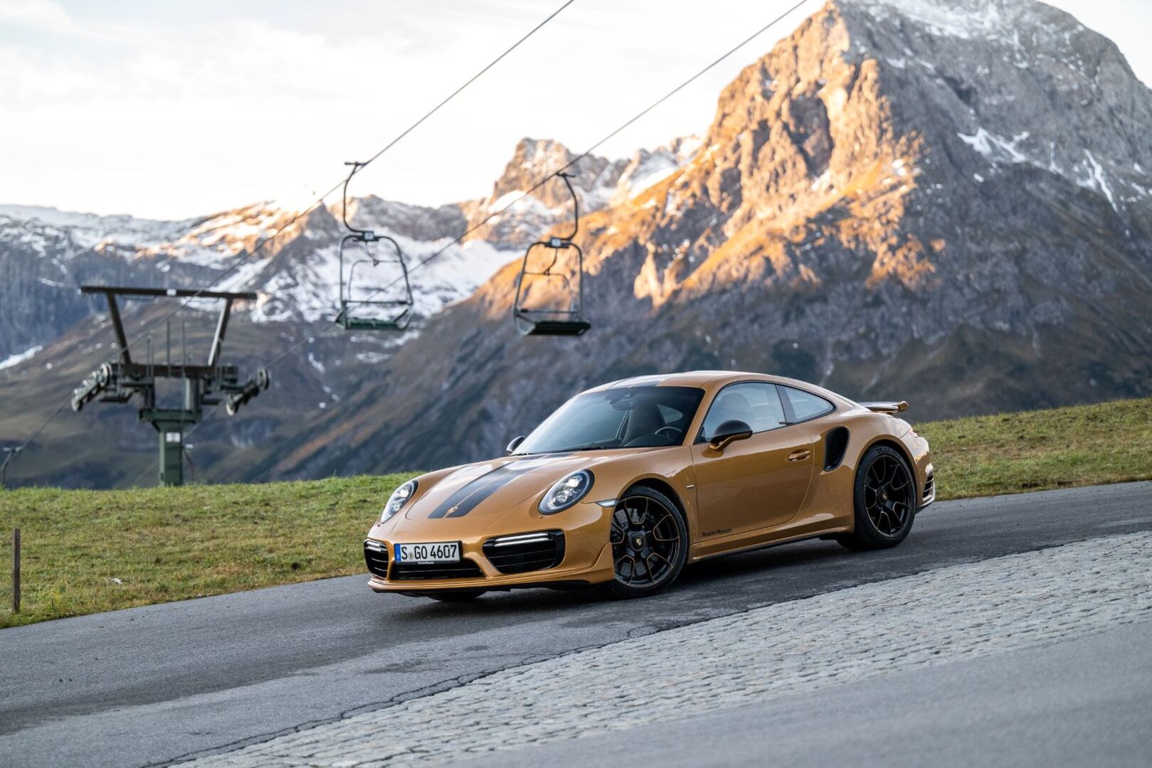 Ein goldener Porsche fährt auf einer schmalen Straße. Im Hintergrund sind schneebedeckte Gipfel und ein Sessellift zu sehen.