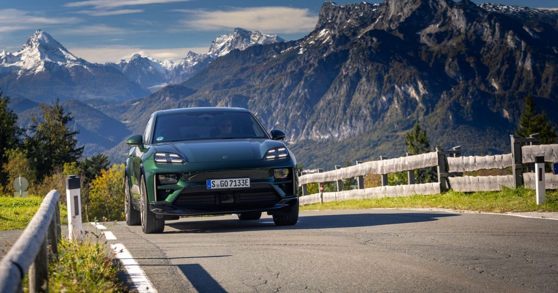 Ein dunkelgrüne Porsche Macan Turbo fährt eine Bergstraße - die von Holzzäunen begrenzt ist - hinauf. Im Hintergrund sind schneebedeckte Gipfel zu sehen.