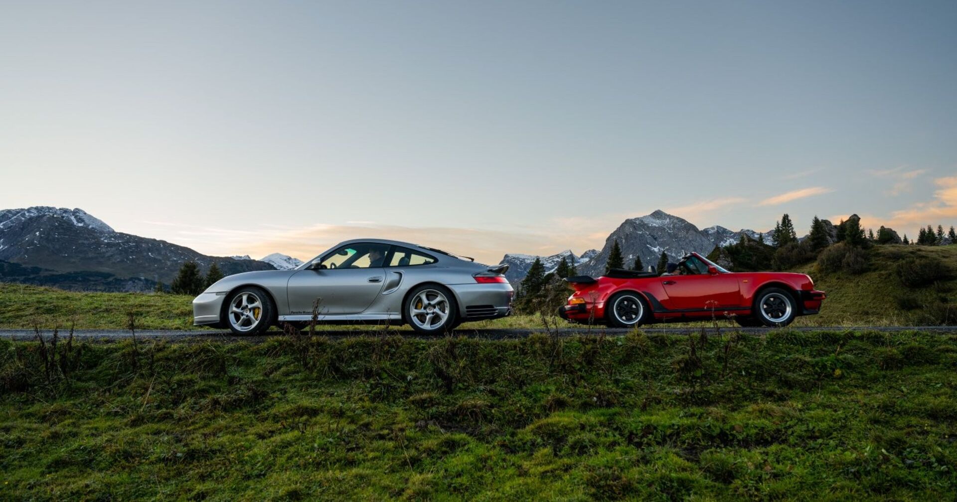 Ein silberner und ein roter Porsche (ein Cabrio) fahren auf einer Bergstraßen. Kurven sind keine zu erkennen, dafür am Horizont schneebedeckte Berge, Wiesen und Bäume.