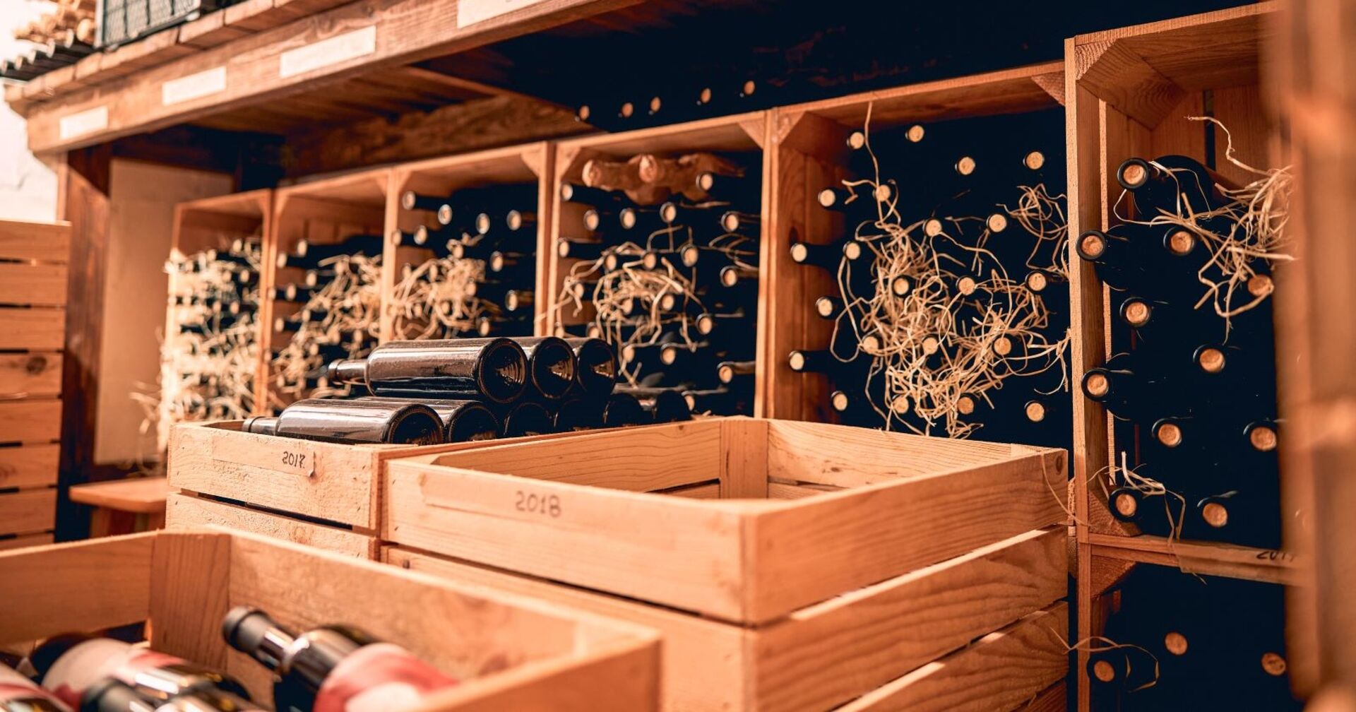 In einem Kellerabteil stehen mit Weinflaschen gefüllte braune Holzkisten und Regale. Einige Weinflaschen sind mit Holzwolle geschützt.