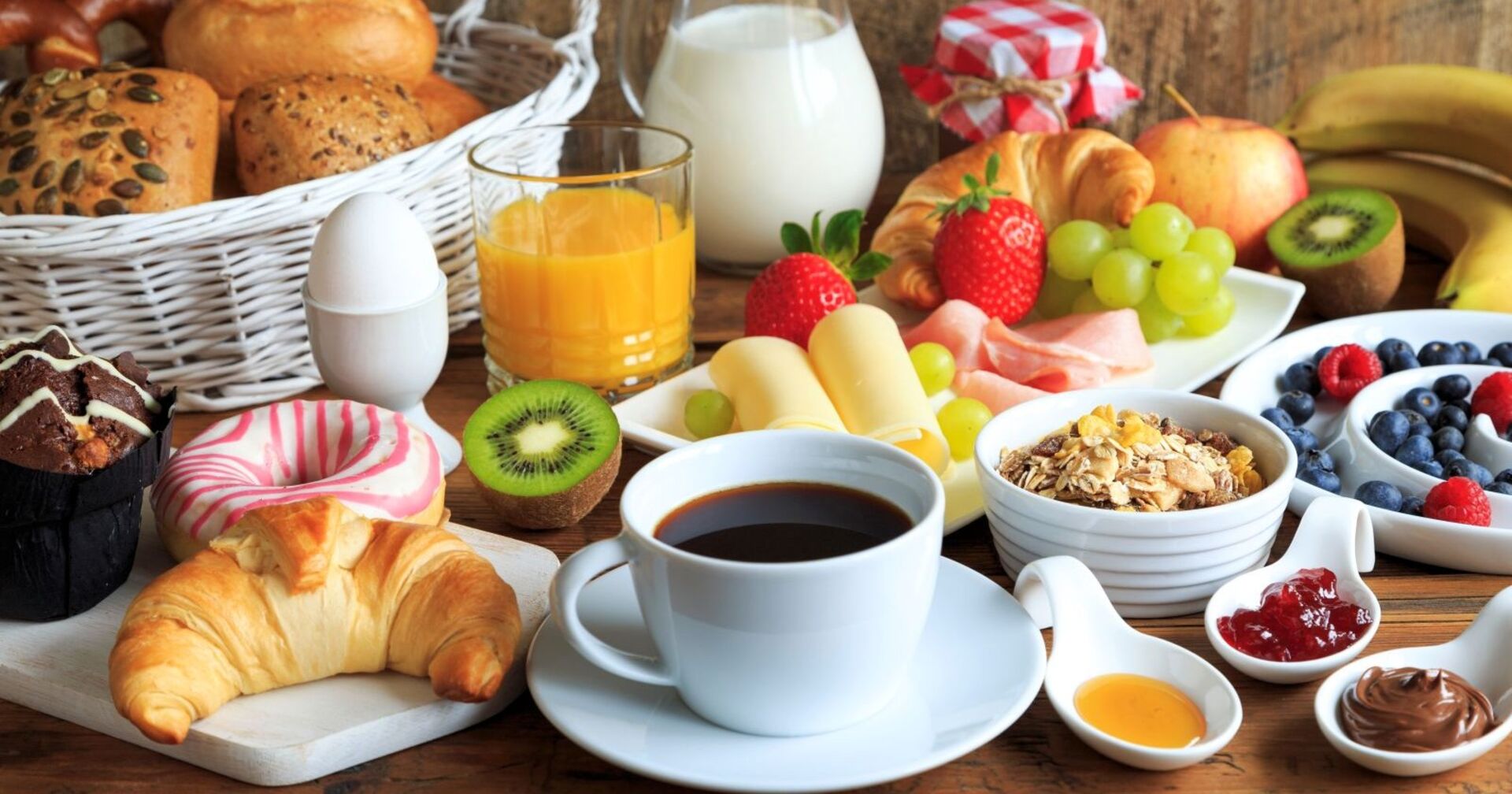 Ein reich gedeckter Frühstückstisch: Zu sehen sind unter anderem eine Tasse Kaffee, Wurst, Käse, ein Korb mit Brötchen, Marmelade, Kiwis, Erdbeeren und andere Früchte.