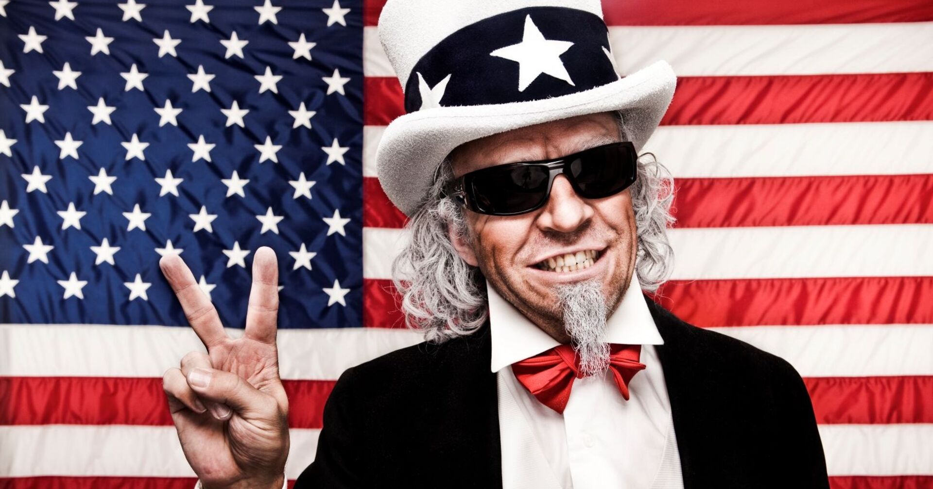 Ein Mann, Symbolfigur Uncle Sam, mit weißem Bart ist zeigt mit seiner rechten Hand das Victory-Zeichen. Er trägt eine schwarze Brille und eine weißen Hut mit schwarzen Band und weißen Sternen. Im Hintergrund ist die Flagge der USA zu sehen.