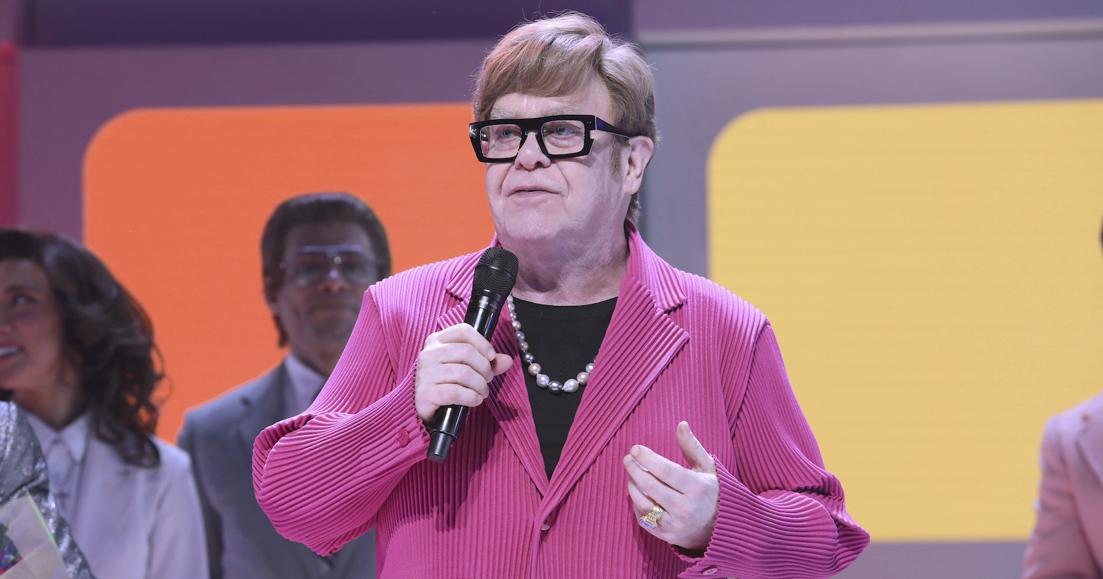 Elton John in einem rosa Sakko auf der Bühne.