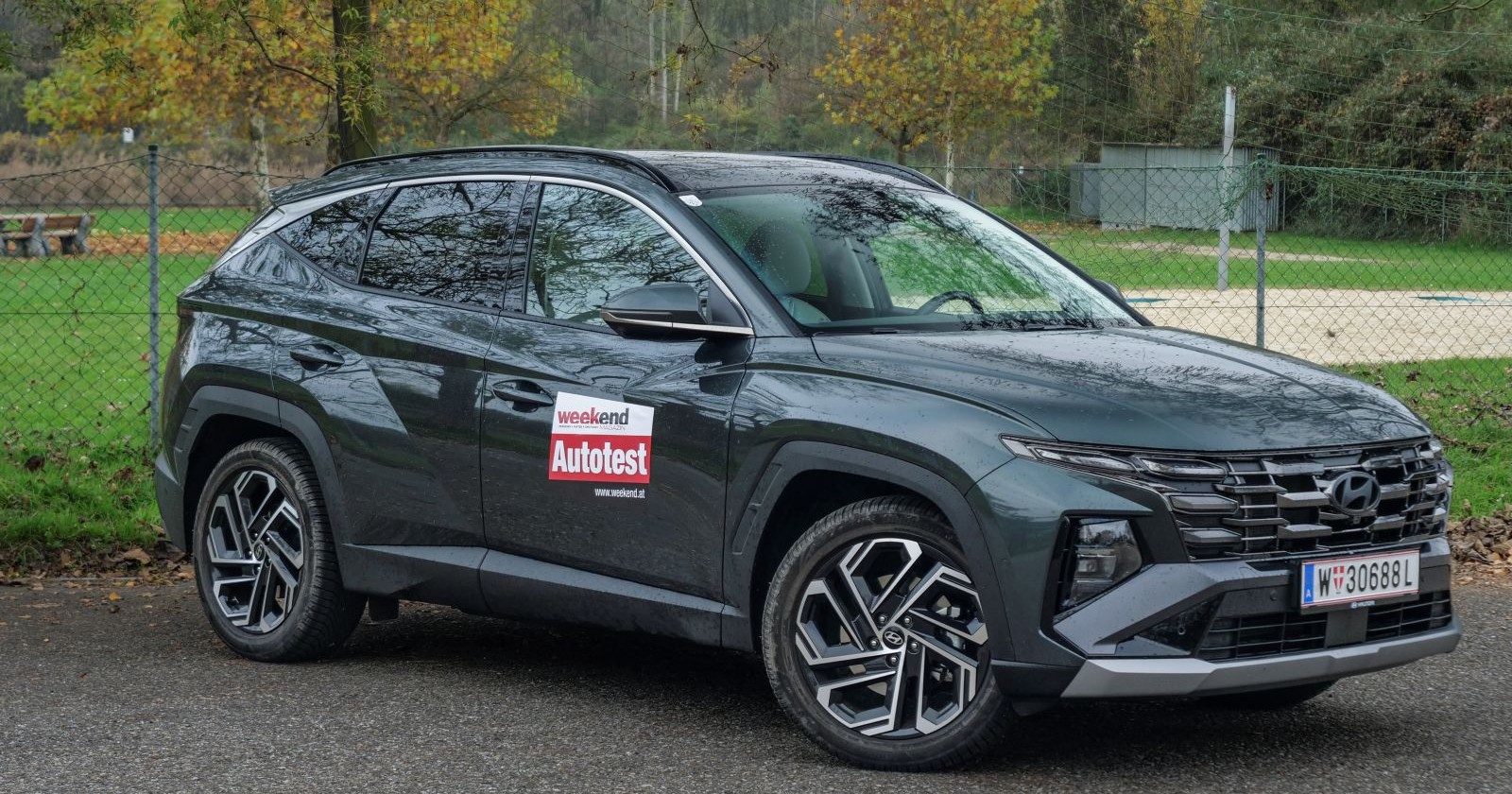 Ein blau-grüner Hyundai Tuscon parkt vor einem Zaun. Im Hintergrund ist eine Wiese, herbstliche Bäume und ein Beachvolley-Platz zu sehen.