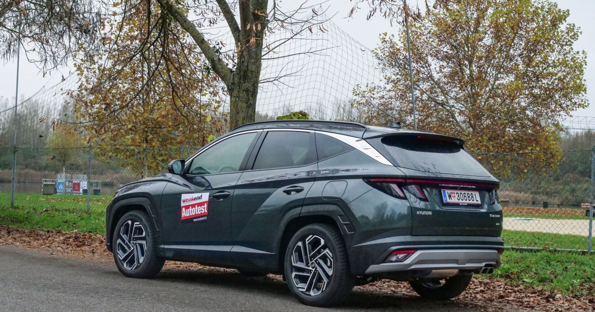 Ein blau-grüner Hyundai Tuscon parkt seitlich an einem Zaun. Im Hintergrund ist eine Wiese, herbstliche Bäume und eine kleine Sandfläche zu sehen.