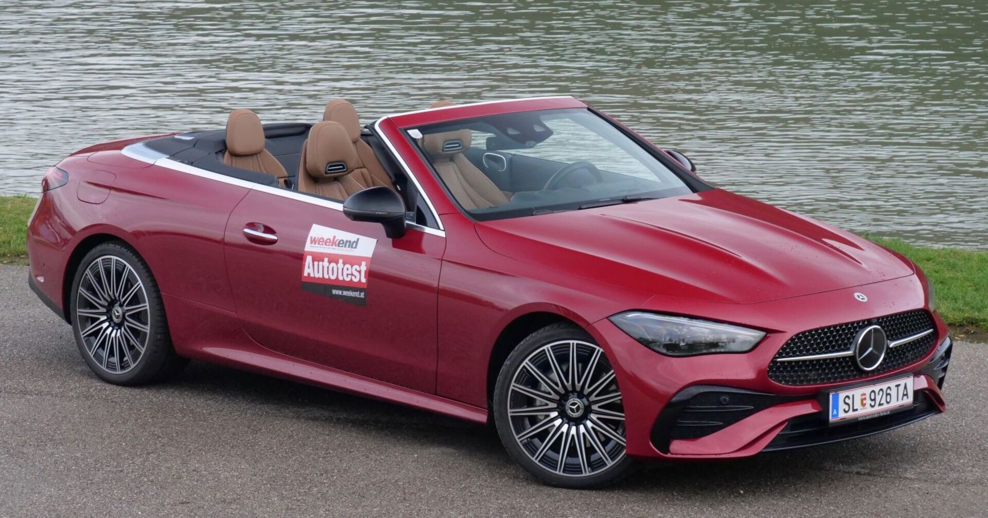 Ein roter Mercedes CLE steht am Rande eines Gewässers. Das Cabrio hat das Verdeck geöffnet. man sieht die braunen Sitze des Autos.