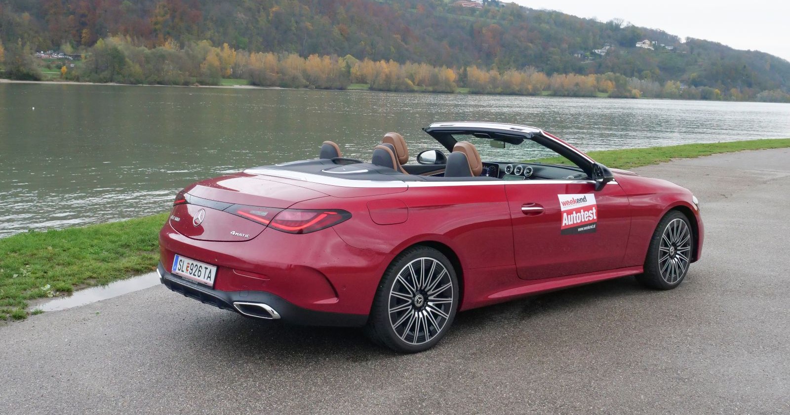 Ein roter Mercedes CLE Cabrio von der Seite. Das Cabrio parkt auf einem asphaltierten Parkplatz, im Hintergrund ist ein Fluss und bewaldete Hügel zu sehen. Das Cabrio hat das Verdeck geöffnet.