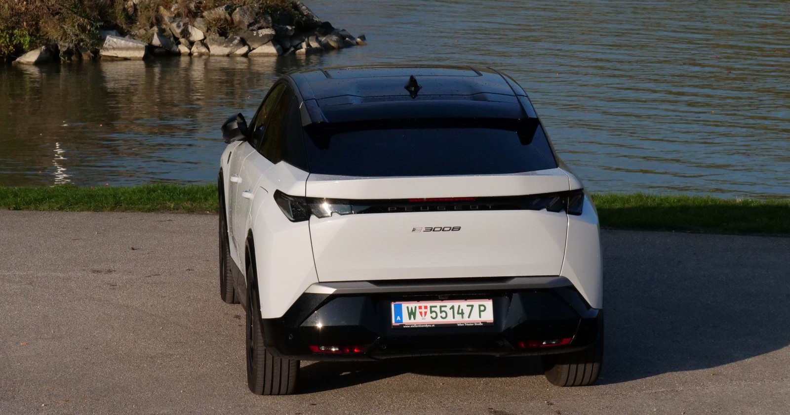 Ein weißer Peugeot E-2008 parkt am Ufer eines Gewässers. Zu sehen ist das Heck des Franzosen, das in der Sonne leuchtet.