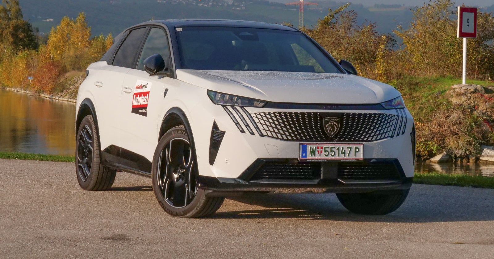 Ein weißer Peugeot E-3008 steht vor einem Gewässer. Die Sträucher im Hintergrund schimmern in den Farben des Herbstes in der Sonne.