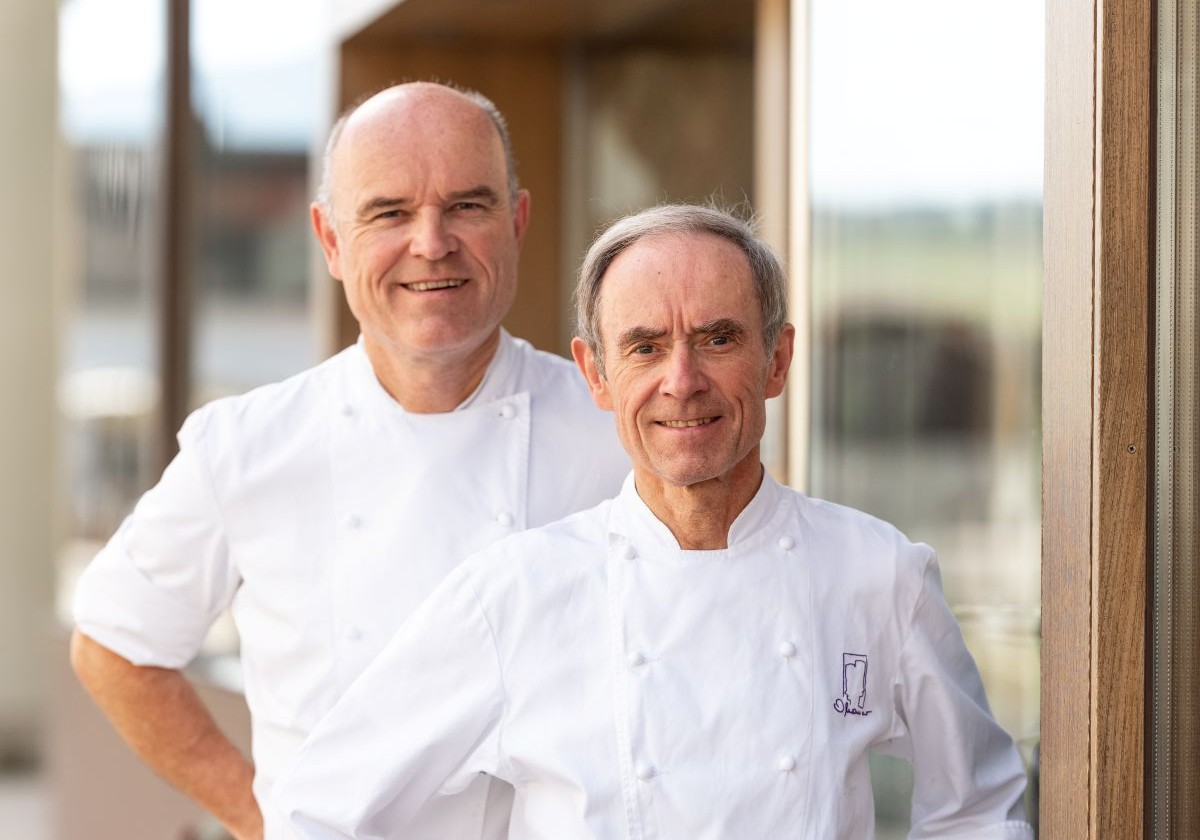 Die beiden Köche Rudolf und Karl Obauer: Beide lachen in die Kamera und tragen weiße Kochjacken. Rudolf Obauer ist größer als sein Bruder, dafür hat er eine Glatze, die bei Karl noch nicht so ausgeprägt ist.