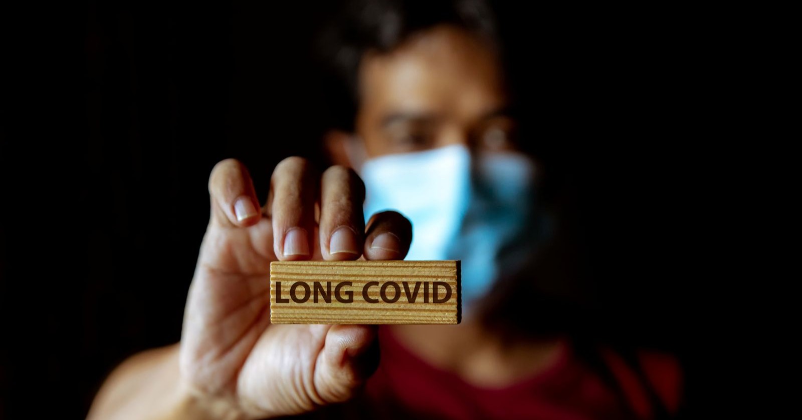 Mann trägt Corona-Maske und hält Holzstück mit Aufschrift &quot;Long Covid&quot; in die Kamera