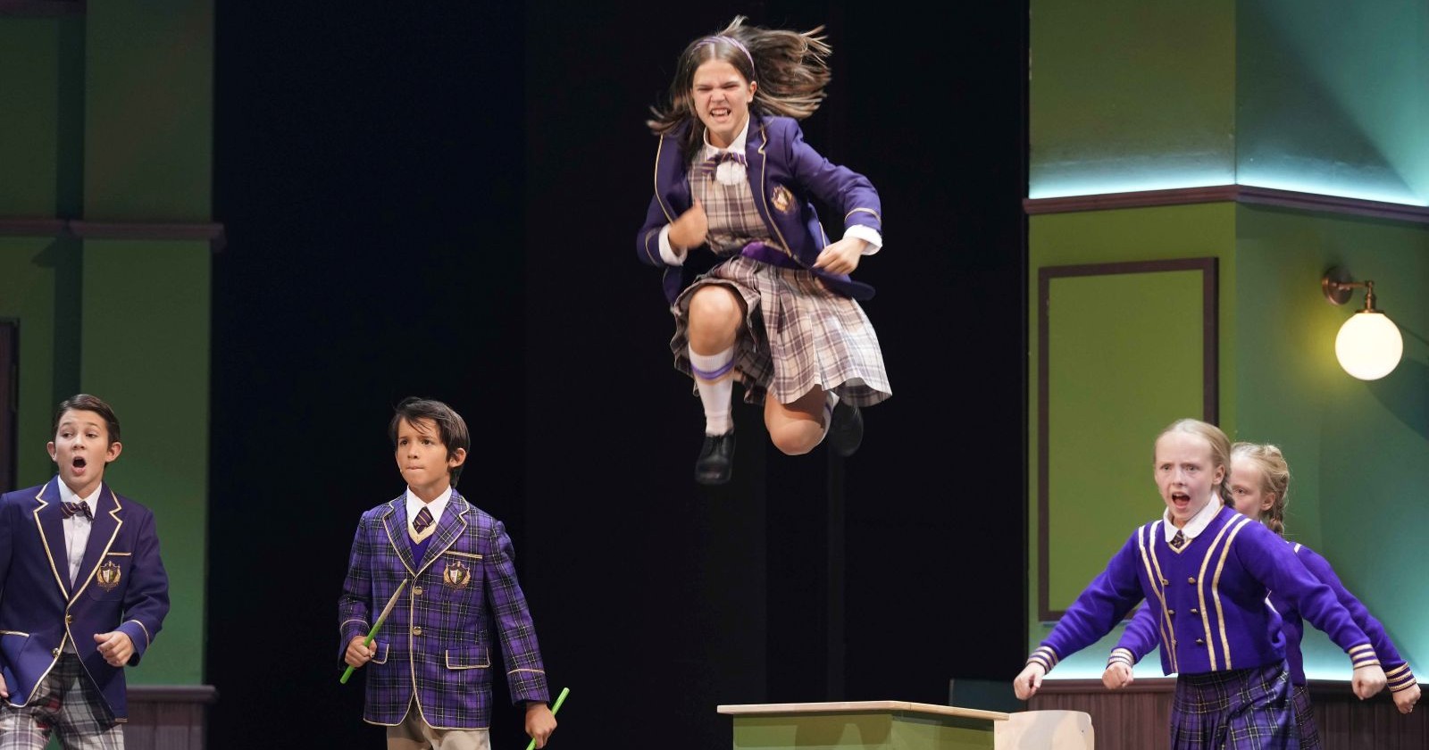 Mädchen und Jungen in Schuluniform singen in einer Klasse. In der Mitte springt ein Mädchen in die Luft. Sie scheint wütend oder besonders engagiert zu sein.