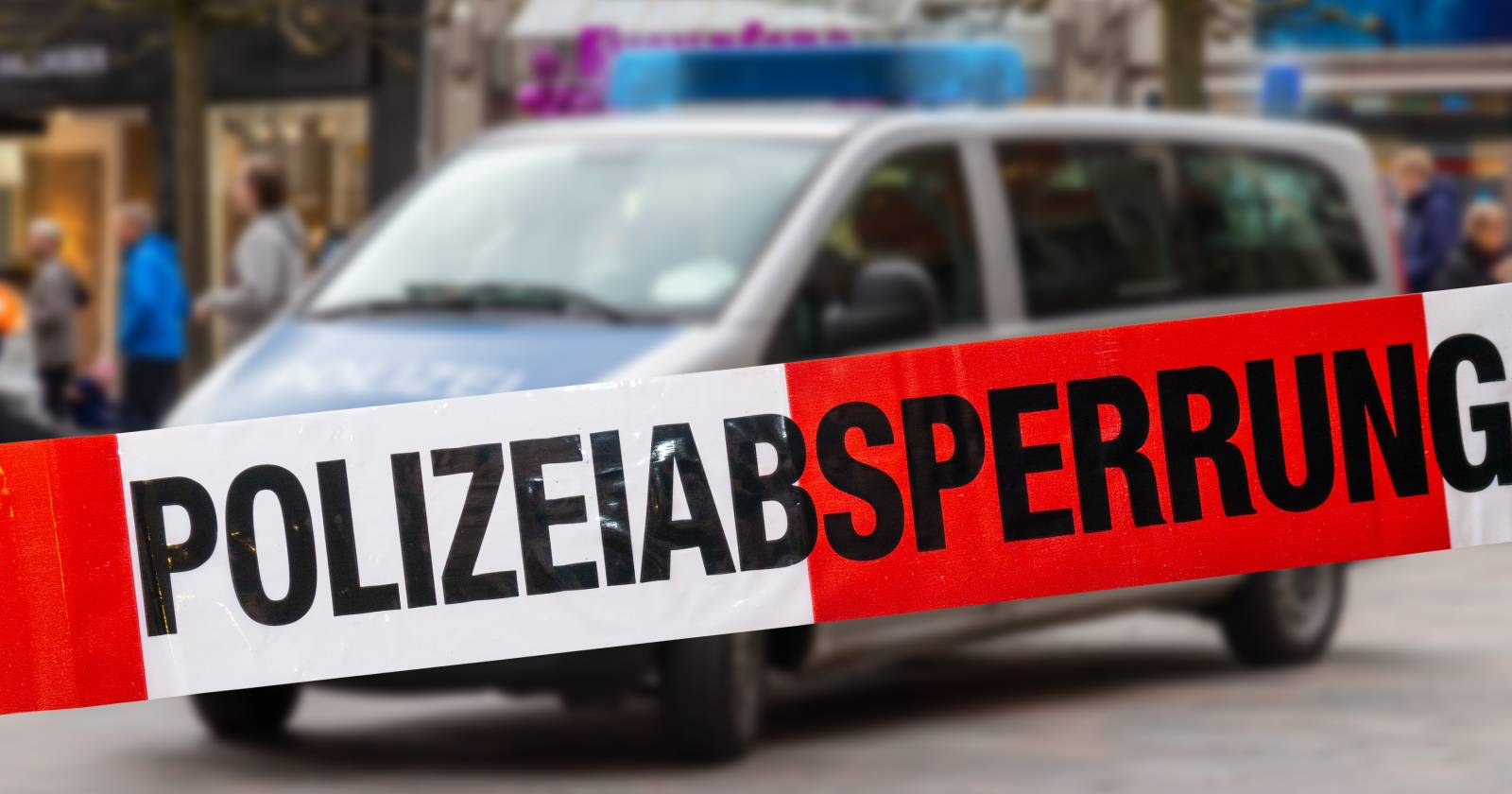 Ein Absperrband auf dem der Schriftzug &quot;Polizeiabsperrung&quot; zu lesen ist und in dessen Hintergrund sich ein leicht verschwommen erscheinendes Polizeiauto befindet