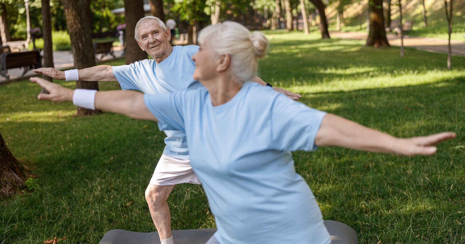 Ein Senior und eine Senioren betreiben Tai-Chi im Park, beide lächeln, während sie eine Übung ausführen.