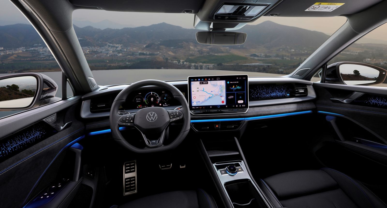 Das Cockpit des VW Tayron ist in grau gehalten. Rechts neben dem Lenkrad ist der Touchscreen zu sehen. Der Blick aus der Windschutzscheibe zeigt ein Gewässer und im Hintergrund sind Berge und Wiesen zu sehen.