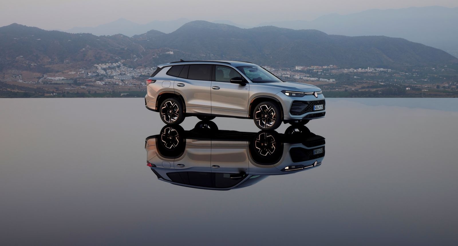 Ein grauer VW Tayron steht auf eine spiegelglatten Fläche, die an einen See erinnert. Im Hintergrund sind kaum bewaldete Berge und in der Ferne einige Häuser zu sehen.