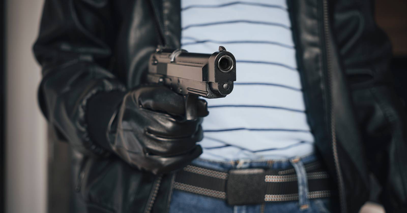 Nahaufnahme des Oberkörpers eines Mannes, der eine schwarze Jacke und schwarze Lederhandschuhe trägt, und eine gezückte Pistole in der Hand hält