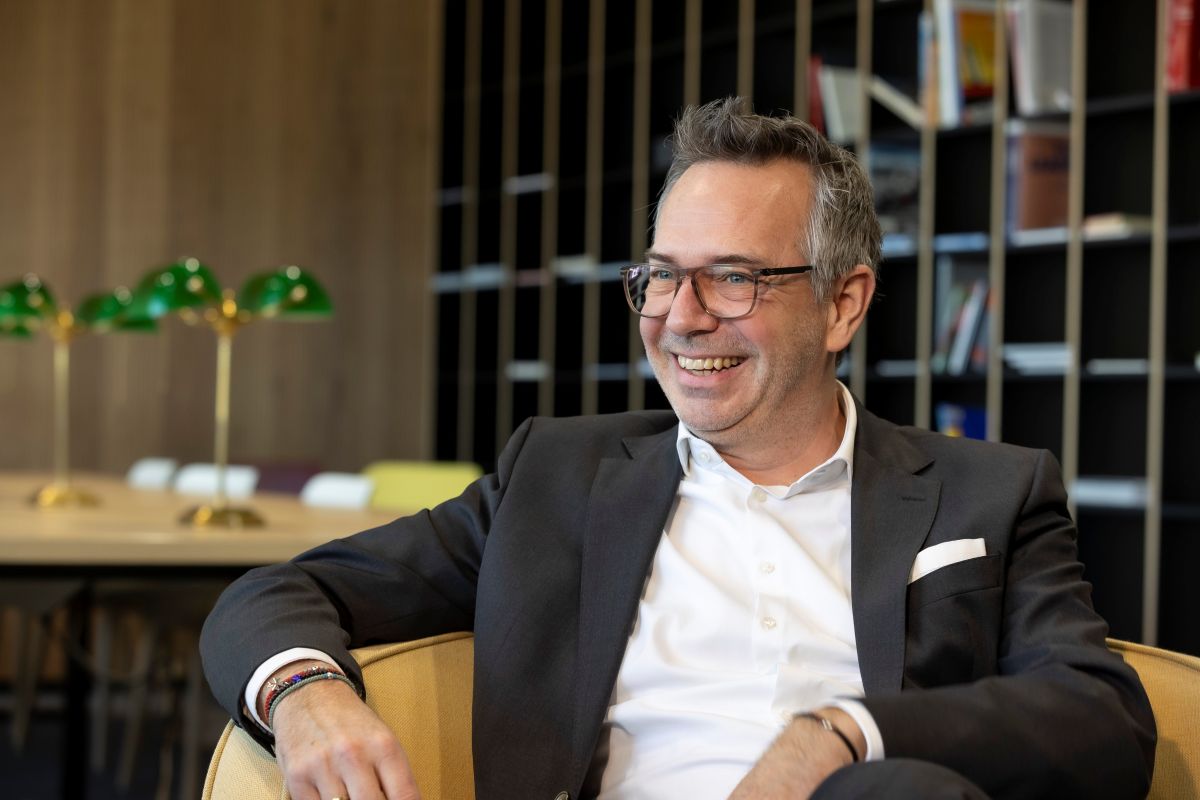 Alexander Bamberger trägt er eine Brille und lacht. Er ist mit einem weißen Hemd und einem schwarzem Sakko bekleidet. Der BMW-Manger sitzt einem braunen Ledersessel. Im Hintergrund ist ein Schreibtisch - auf dem grüne Lampen stehen - zu sehen. Weiters im Bild: Ein Regal in dem Zeitungen und Bücher sortiert sind.