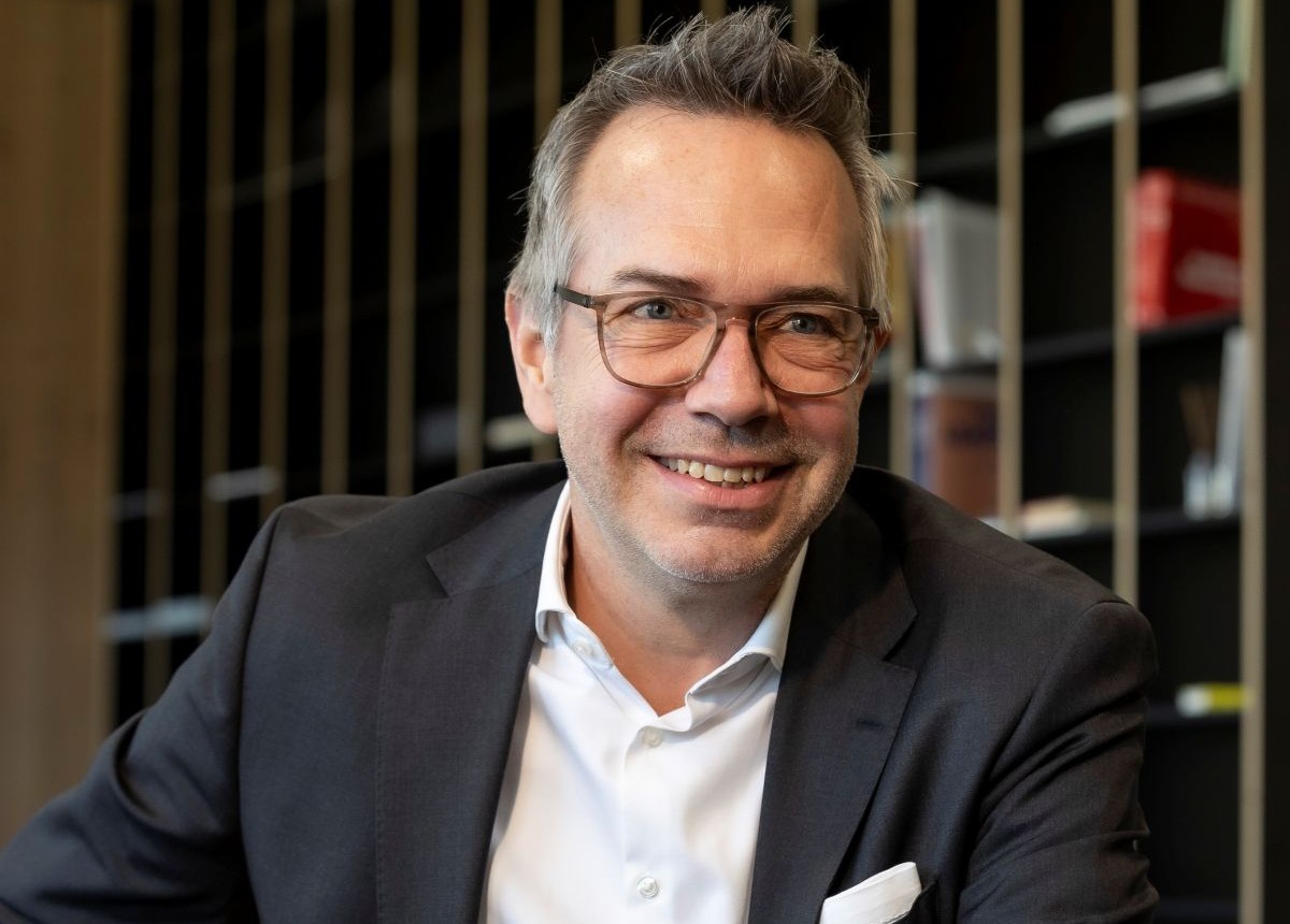 Alexander Bamberger trägt er eine Brille und lacht. Er ist mit einem weißen Hemd und einem schwarzem Sakko bekleidet.