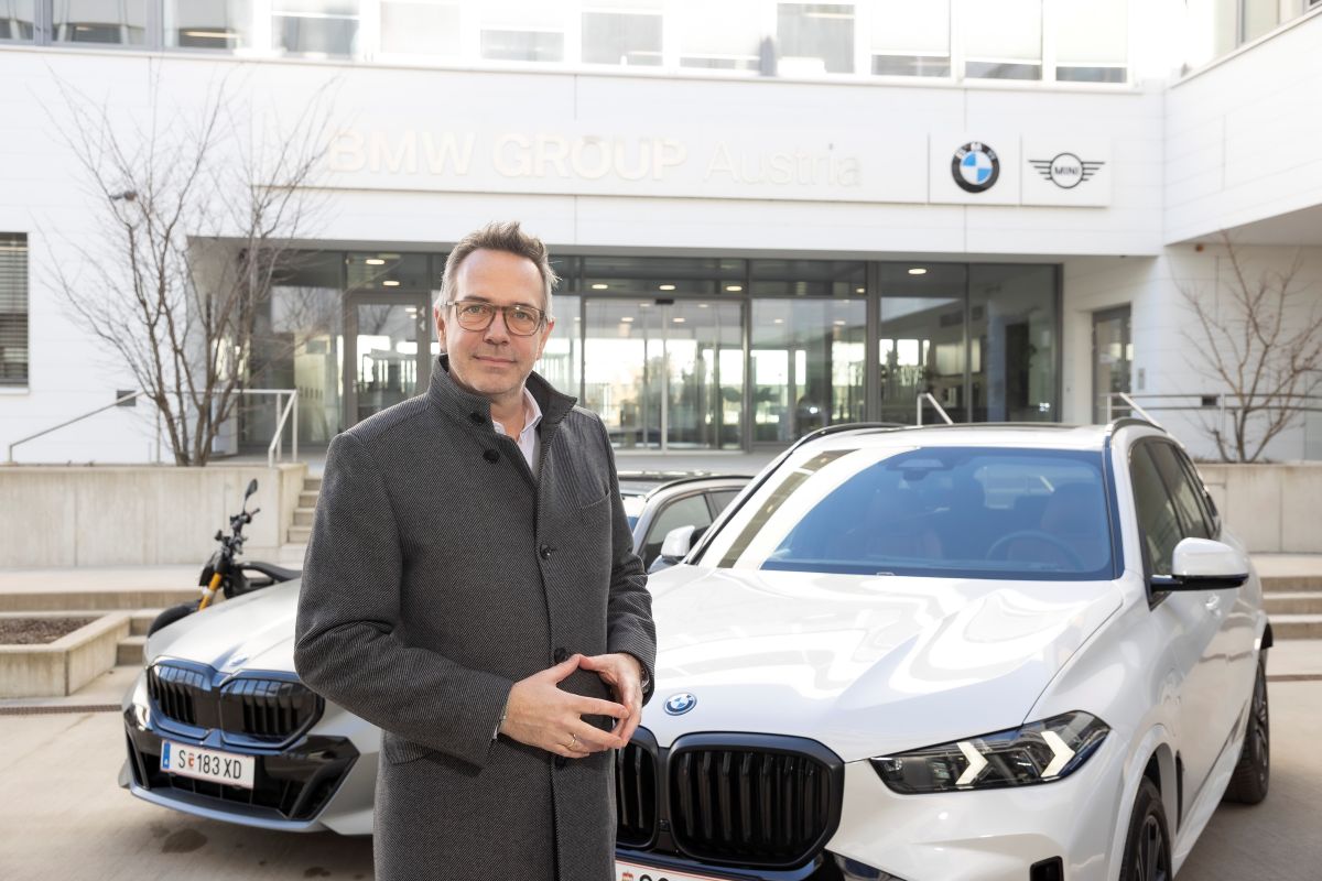 Alexander Bamberger, trägt eine Brille und ist mit einem grauen Mantel bekleidet. Der BMW-Manager steht vor zwei weißen BMWs. Im Hintergrund ist ein Gebäude mit einer großen Glasfläche zu sehen. Am Gebäude ist das BMW-Logo angebracht.
