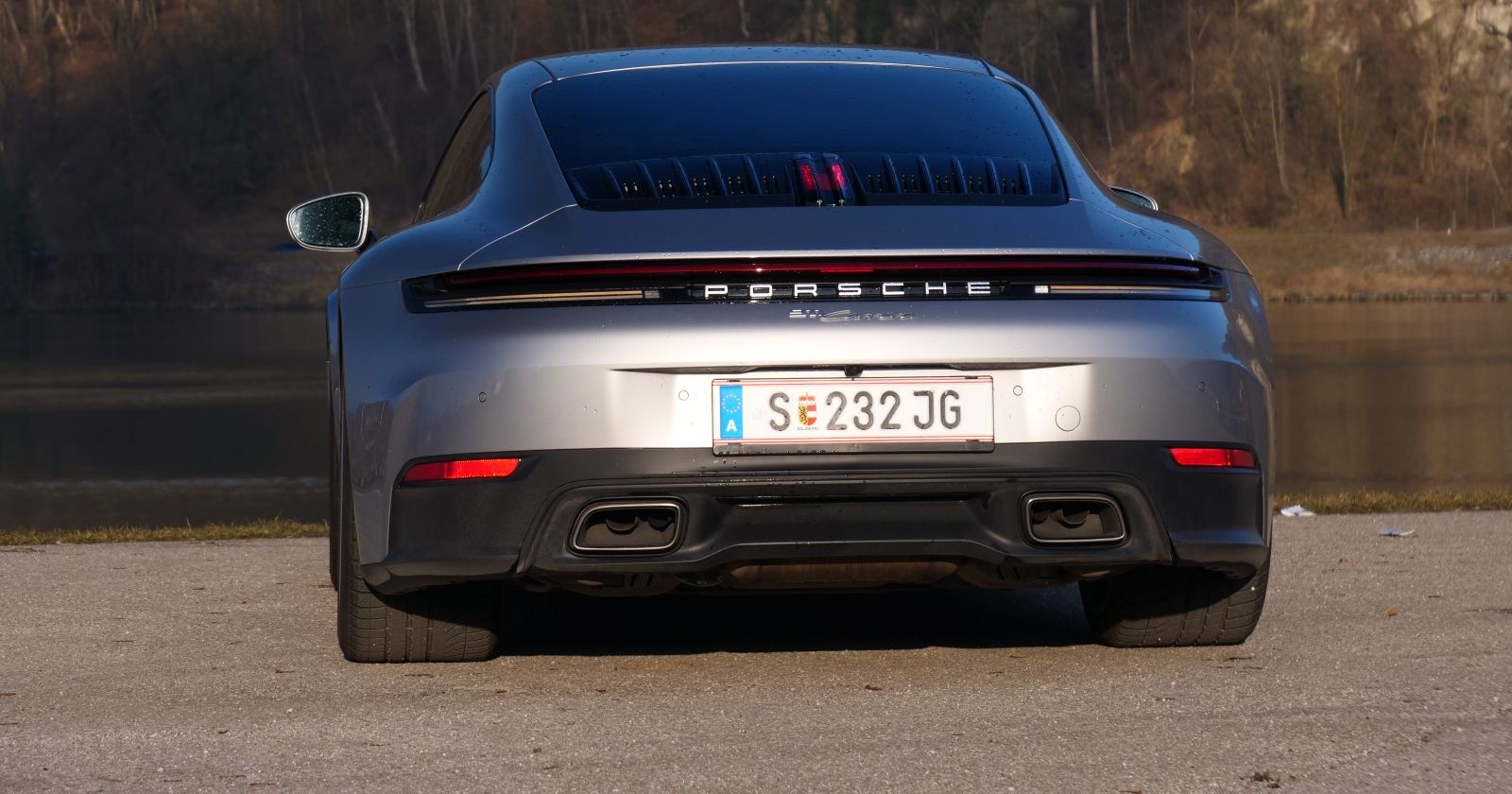 Das silberne Heck des Porsche 911 strahlt in der Sonne. Das Auto steht am Ufer eines Gewässers.