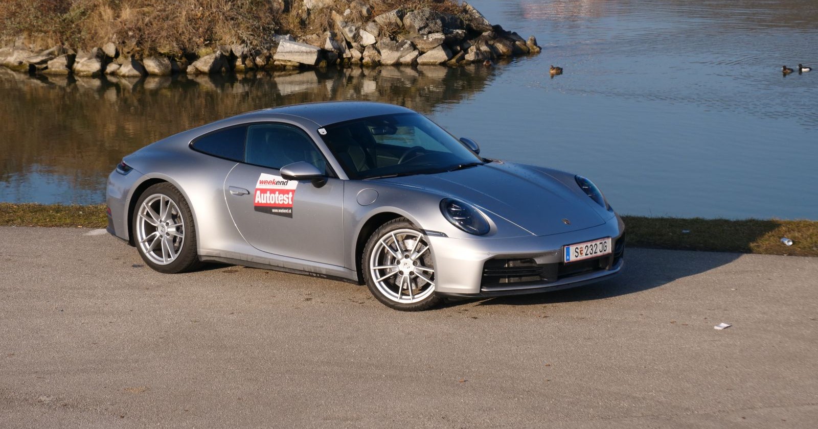 Ein silberner Porsche 911 parkt am Ufer eines Gewässer in dem einige Enten schwimmen. Das Auto leuchtet in der Sonne.