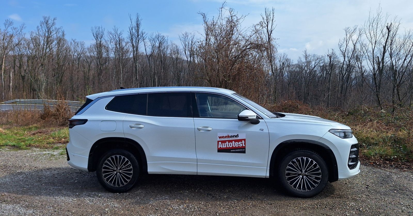 Ein weißer VW Tayron steht auf Schotterparkplatz. Im Hintergrund sind Bäume ohne Blätter zu sehen. Der Himmel ist blau, dennoch sind einige Wolken zu sehen.
