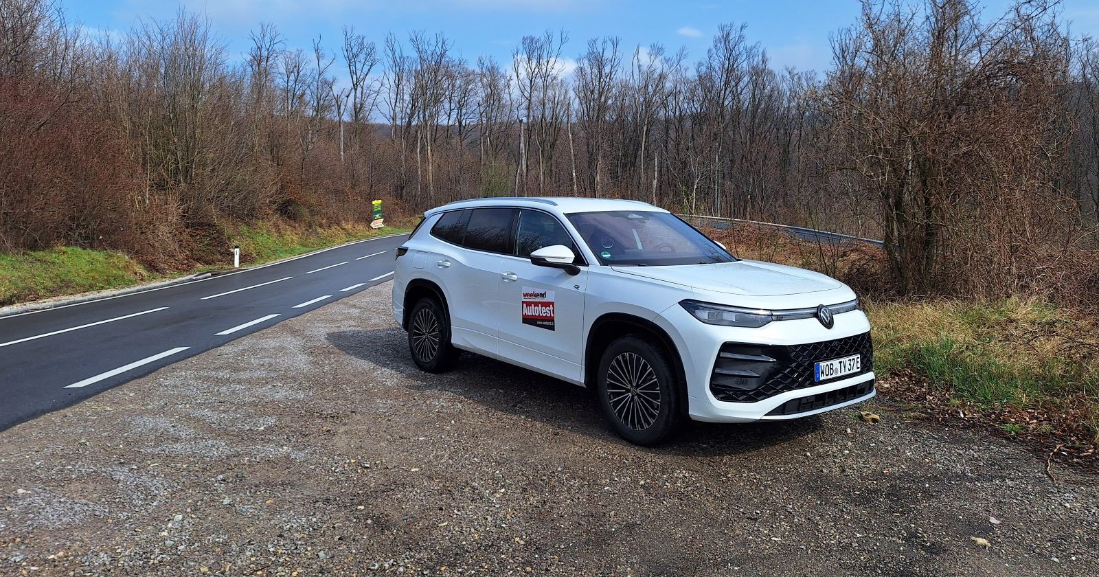 Ein weißer VW Tayron parkt neben einer Landstraße auf der keine Autos unterwegs sind. Am blauen Himmel sind einige Wolken zu sehen, die Bäume in der Landschaft tragen noch keine Blätter.