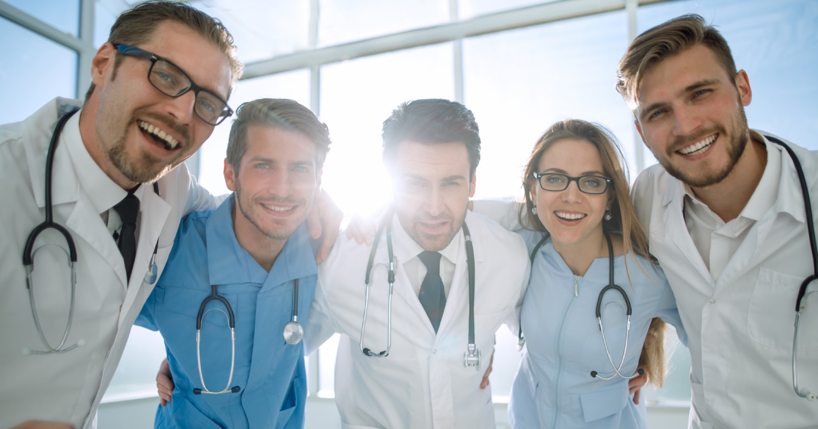 Fünf Ärzte, die einen halbrunden Kreis vor einer sonnendurchfluteten Fensterfront bilden und dabei die Arme über ihren Schultern verschränken