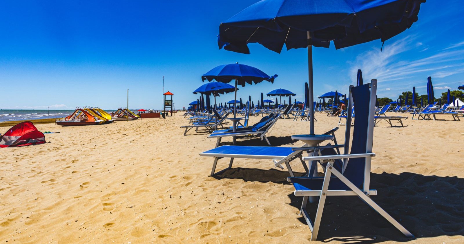 Leerer Sandstrand in Bibione mit aufgestellten, geschlossenen blauen Sonnenschirmen und Liegen.