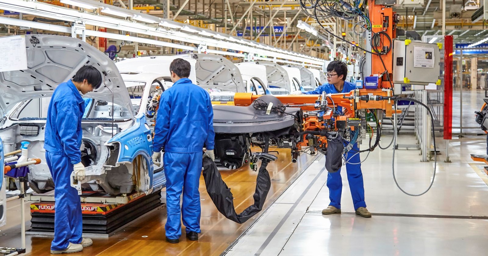 Drei chinesische Arbeiter - bekleidet mit blauen Arbeitsgewand - stehen in einer modernen Fertigungsstraße für Autos.