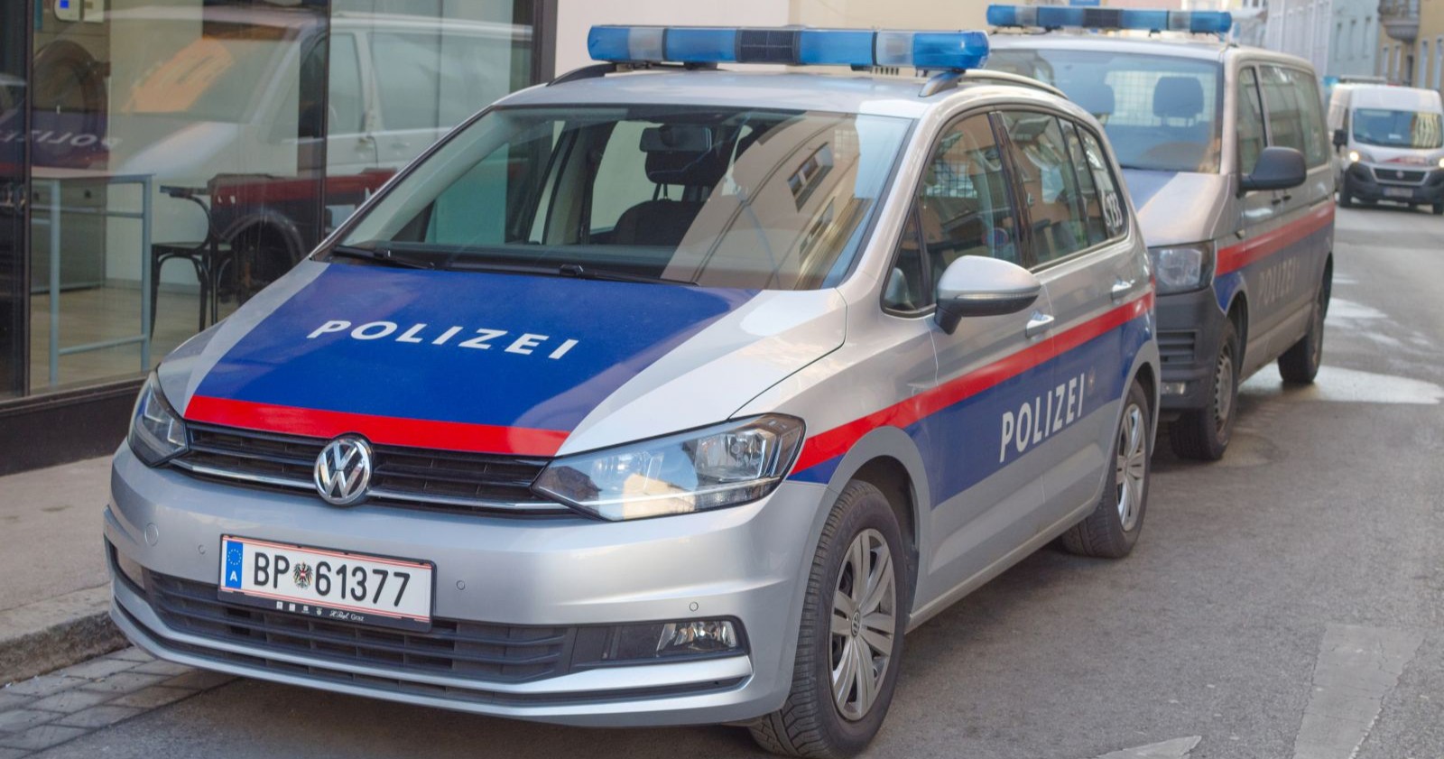 Zwei österreichische Polizeiautos, ein silberner VW mit der Aufschrift &quot;Polizei&quot; in Blau und Rot sowie ein weiteres Einsatzfahrzeug, parken auf einer Straße in der Grazer Innenstadt. Im Hintergrund sind Gebäude und ein Schaufenster zu sehen.