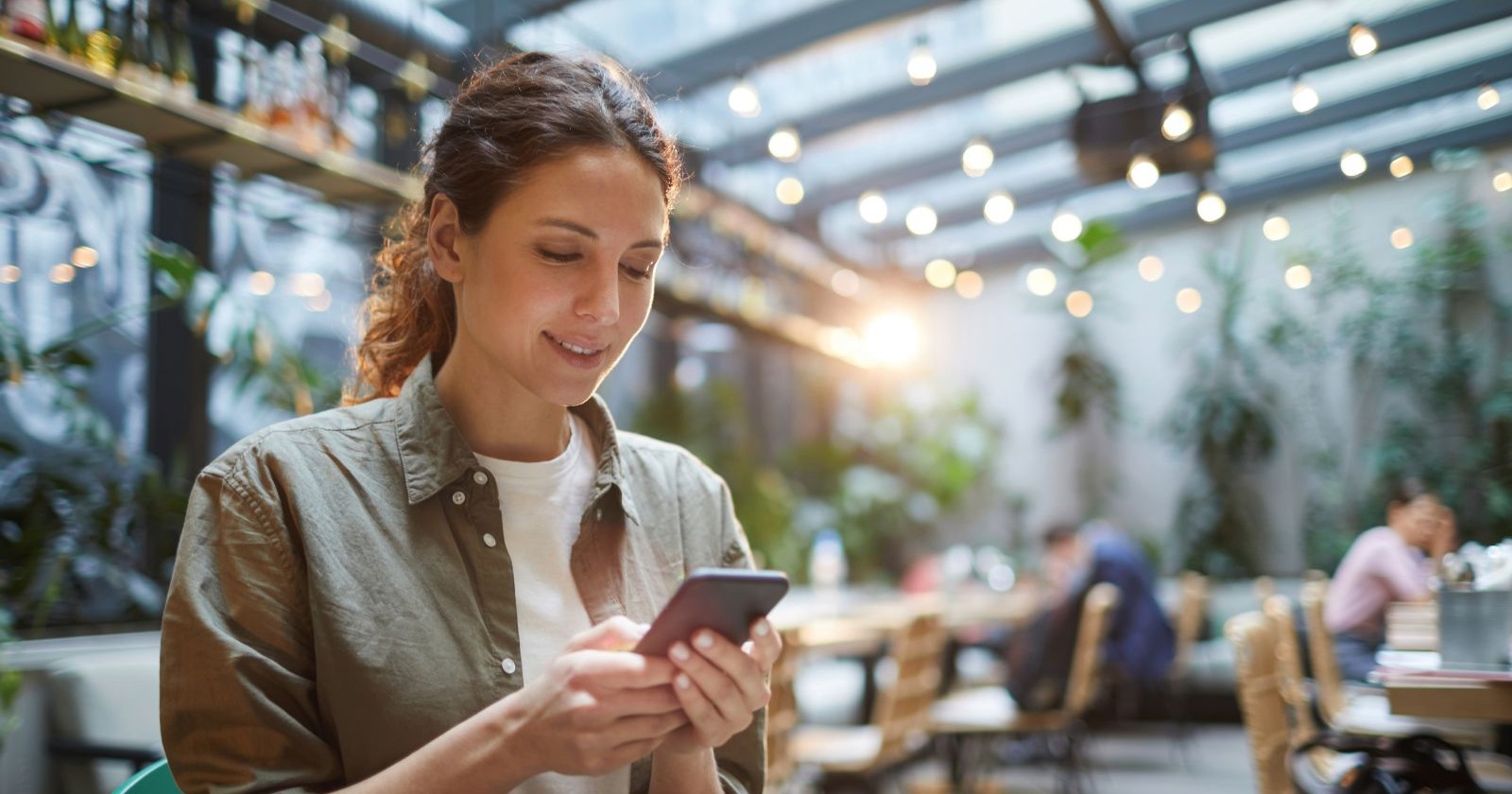 Eine Frau mit braunen Haaren und einem Zopf blickt lächelnd auf ihr Smartphone. Die Frau ist mit einem olivgrünem Hemd und einem weißen T-Shirt bekleidet. Sie sitzt in einem Wintergarten eines Restaurants.