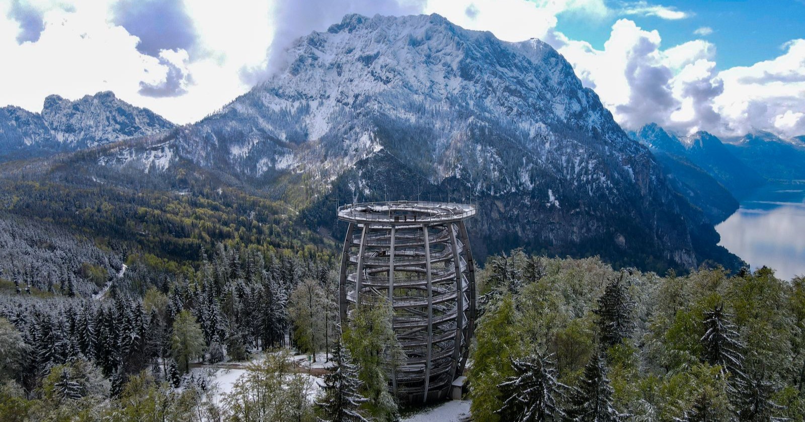 Panoramablick auf den verschneiten Grünberg bei Gmunden mit dem Baumwipfelpfad im Vordergrund und dem Traunstein im Hintergrund. Die umliegenden Wälder sind mit frischem Schnee bedeckt, der Himmel zeigt eine Mischung aus Sonne und dramatischen Wolken.