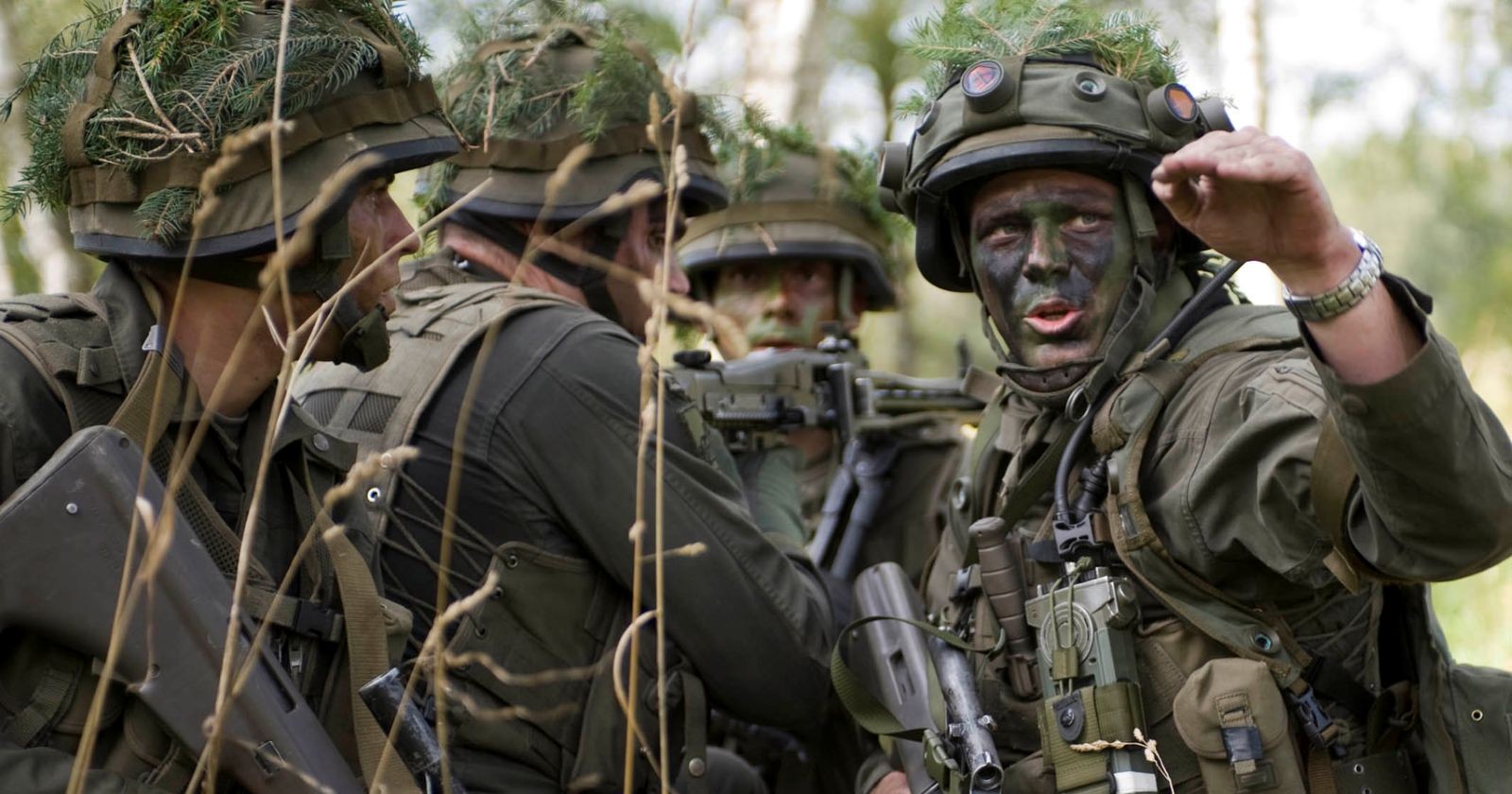 Soldaten des österreichischen Bundesheeres in voller Ausrüstung während einer Übung im Wald. Die Gesichter sind mit Tarnfarbe bemalt, Helme mit Zweigen getarnt. Ein Soldat gibt gestikulierend Anweisungen