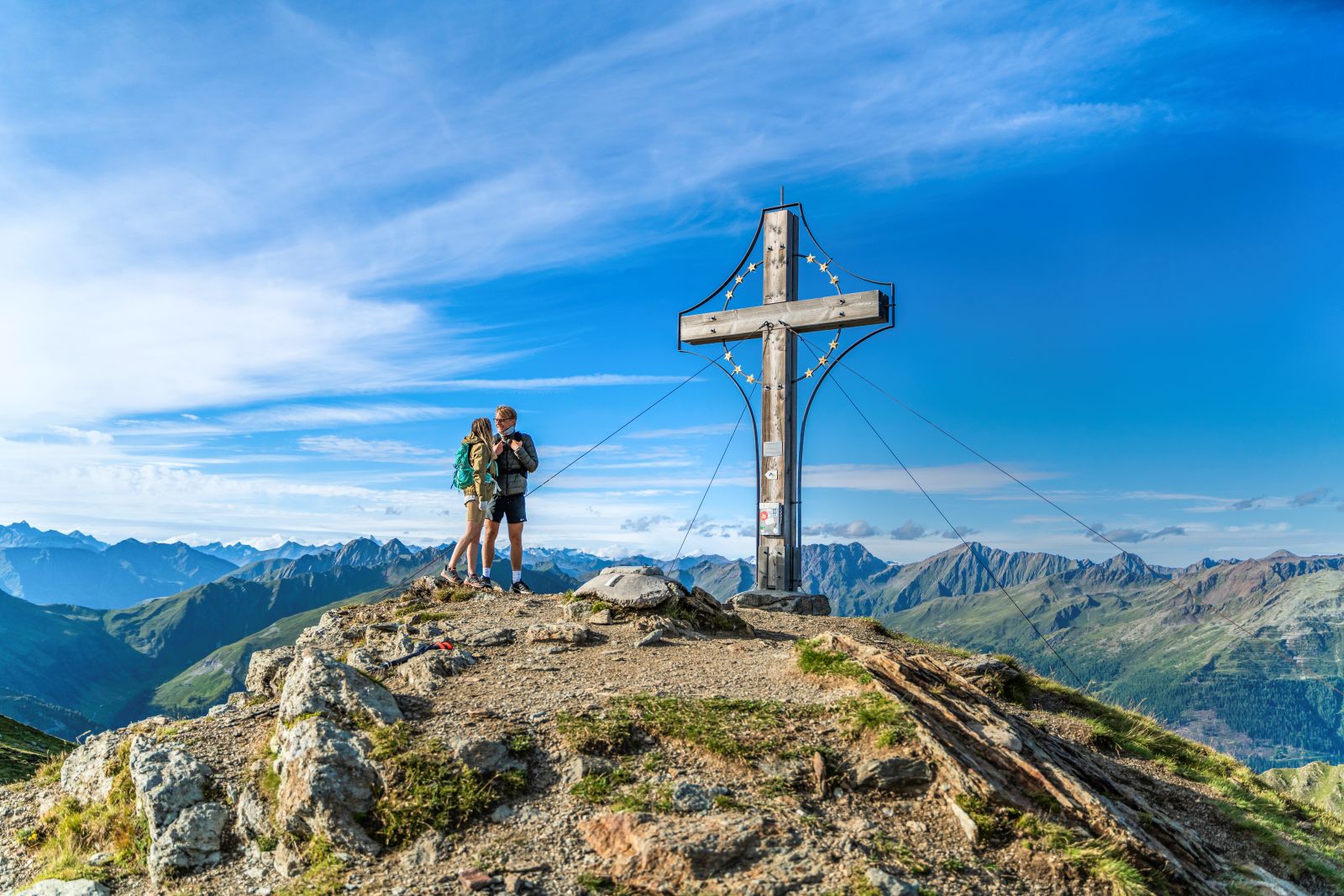 Eine Frau und ein Mann in Wanderoutfit steht am Gipfelkreuz, im Hinterrund Berge und ein strahlend blauer Himmel mit wenigen Wolken.