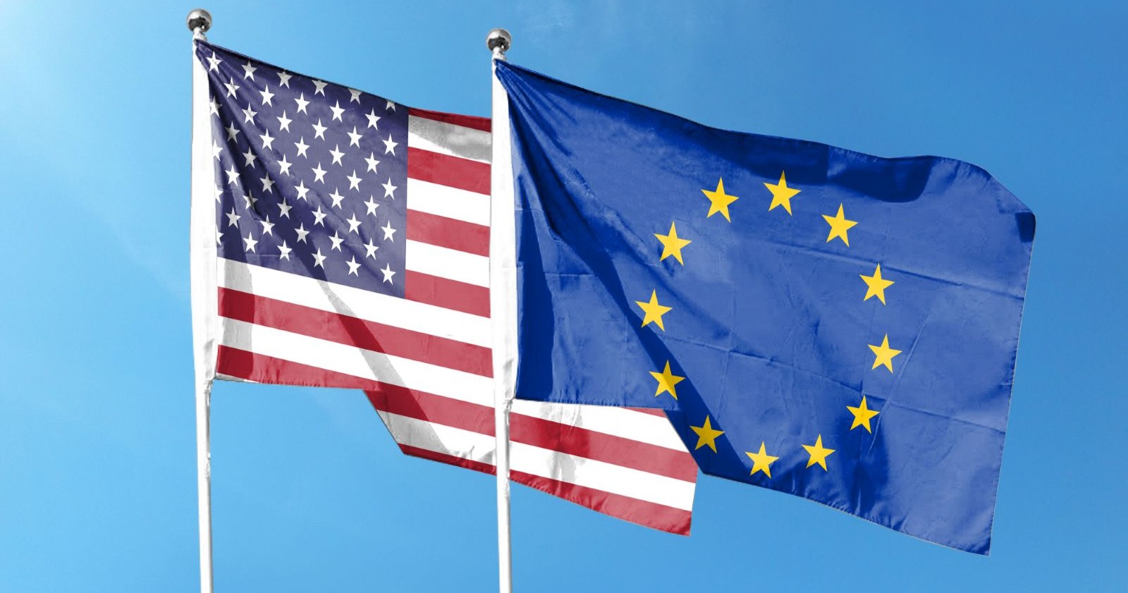 Eine US- und eine EU-Flagge nebeneinander gehisst, im Wind wehend vor blauem Himmel.