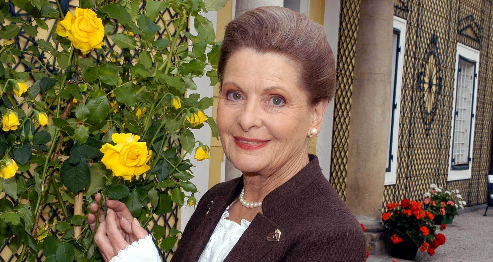 Johanna Matz steht lächelnd neben einem blühenden Rosenstrauch mit gelben Rosen. Sie trägt ein braunes Kostüm mit Perlenkette und blickt freundlich in die Kamera. Im Hintergrund ist ein Gebäude mit kunstvoll verzierten Fenstern und Blumenschmuck zu sehen.