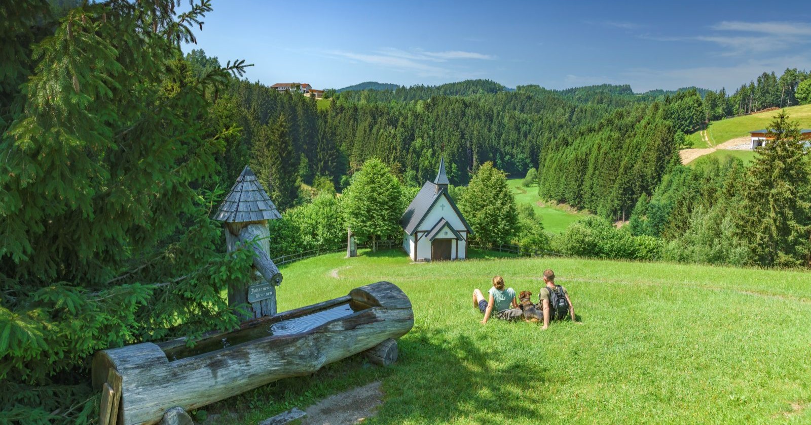 Das Bild zeigt eine sanfthügelige grüne Landschaft, im Vordergrund steht ein Trinkwasserbrunnen aus Holz, in der Mitte sitzt eine Familie auf der Wiese und im Hintergrund steht eine Kapelle.