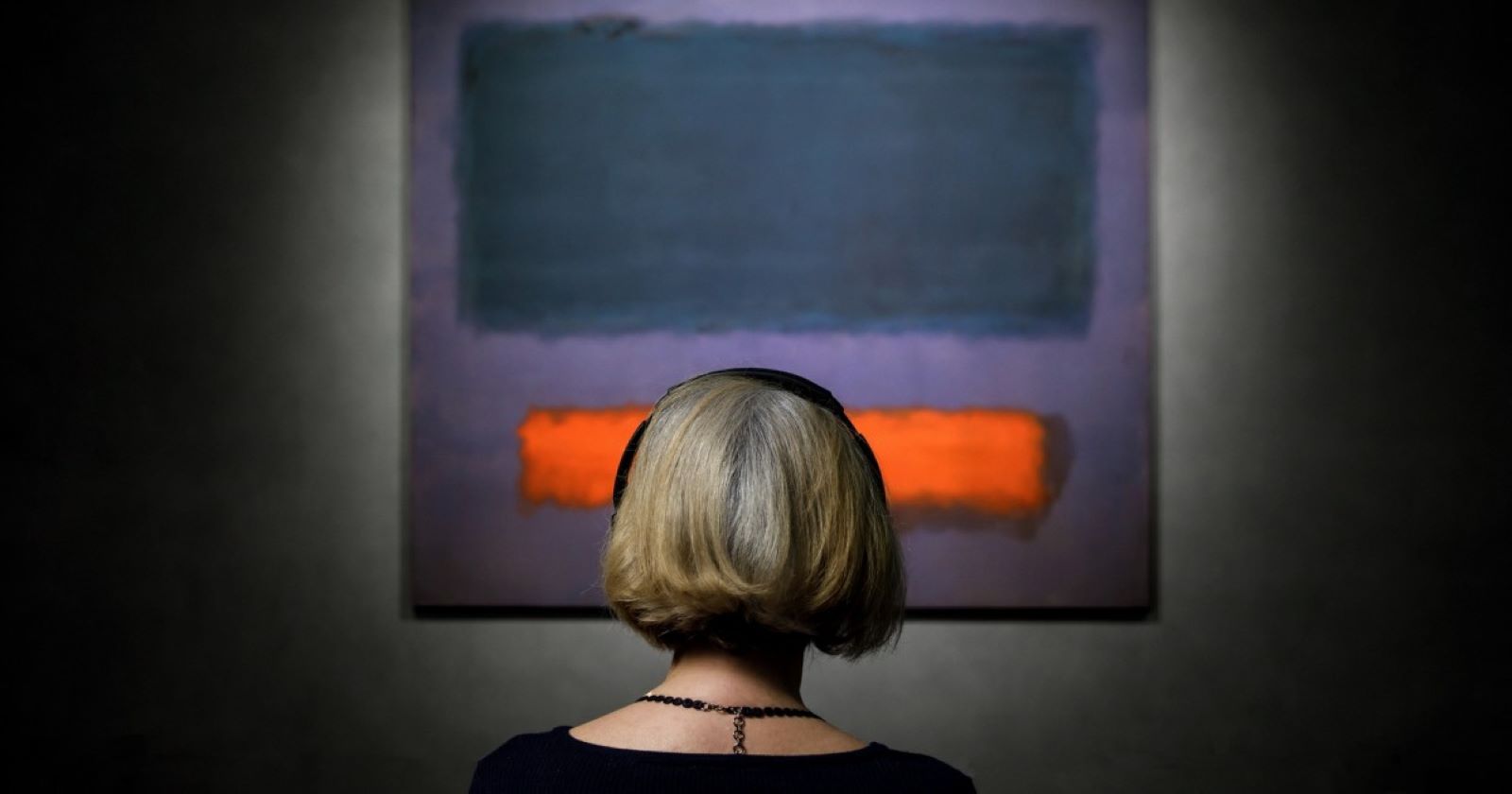 Eine Frau sitzt vor dem - noch unzerkratzten - Gemälde Grey, Orange on Maroon, No. 8 von Mark Rothke. Es zeigt einen dunkelblauen und einen orangen Farblock auf ebenfalls dunkelblauem, aber etwas hellerem Untergrund.