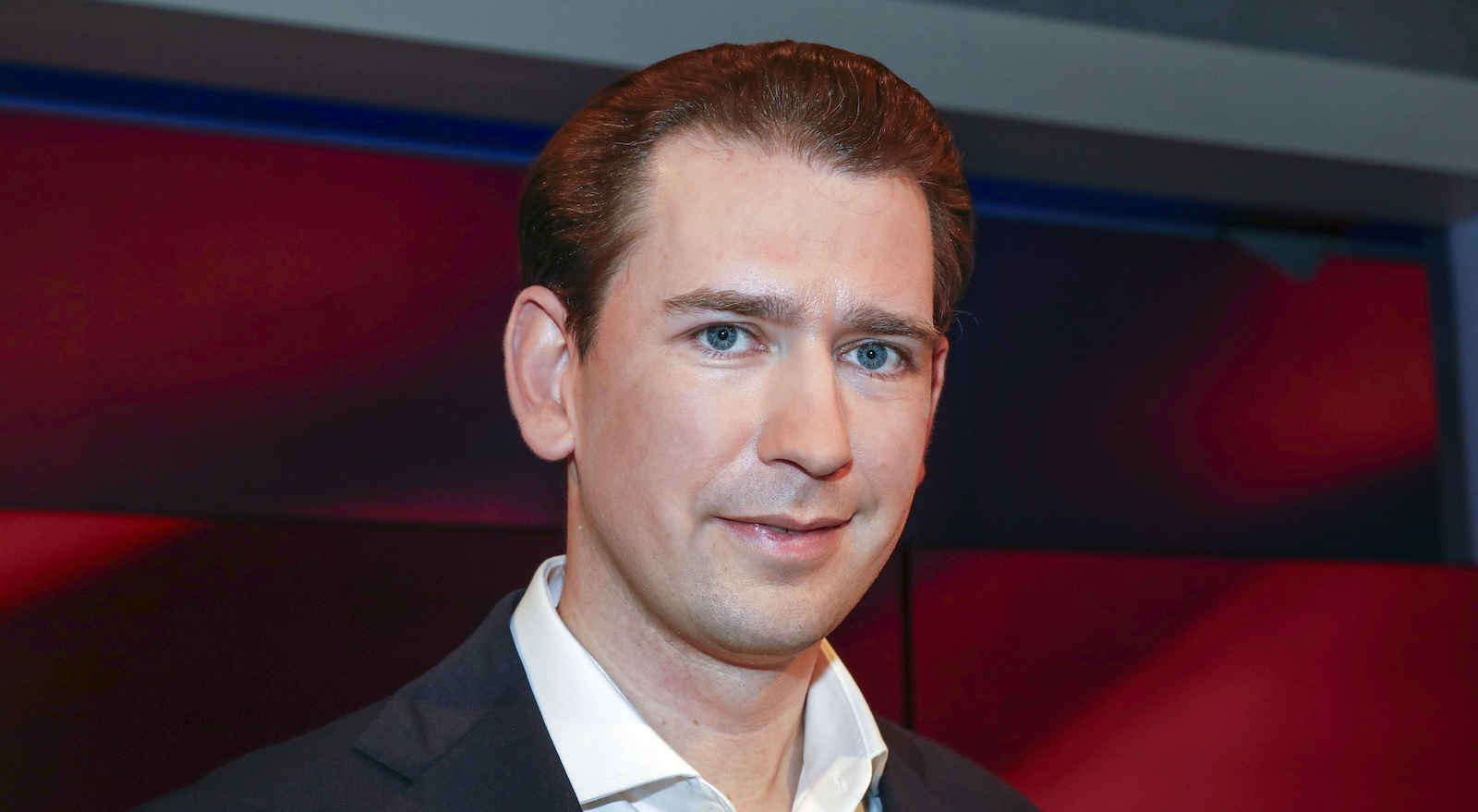 Porträtfoto von Sebastian Kurz in Anzug und weißem Hemd, vor dunklem, rötlich beleuchtetem Hintergrund – Blick in die Kamera, neutraler Gesichtsausdruck.