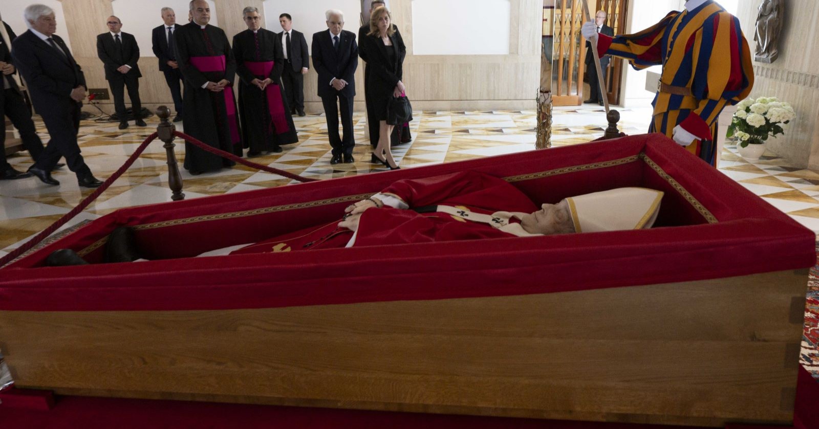 Papst Franziskus liegt im offenen Sarg in rotem liturgischem Gewand. Um den Sarg versammeln sich Geistliche, Politiker und ein Mitglied der Schweizergarde.
