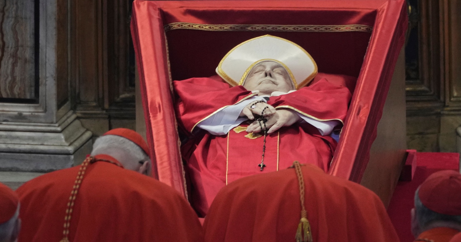 Papst Franziskus liegt im roten Gewand mit Mitra und Rosenkranz in einem geöffneten, schräg gestellten Sarg. Vor dem Sarg stehen mehrere Kardinäle in roter Kleidung und verneigen sich.
