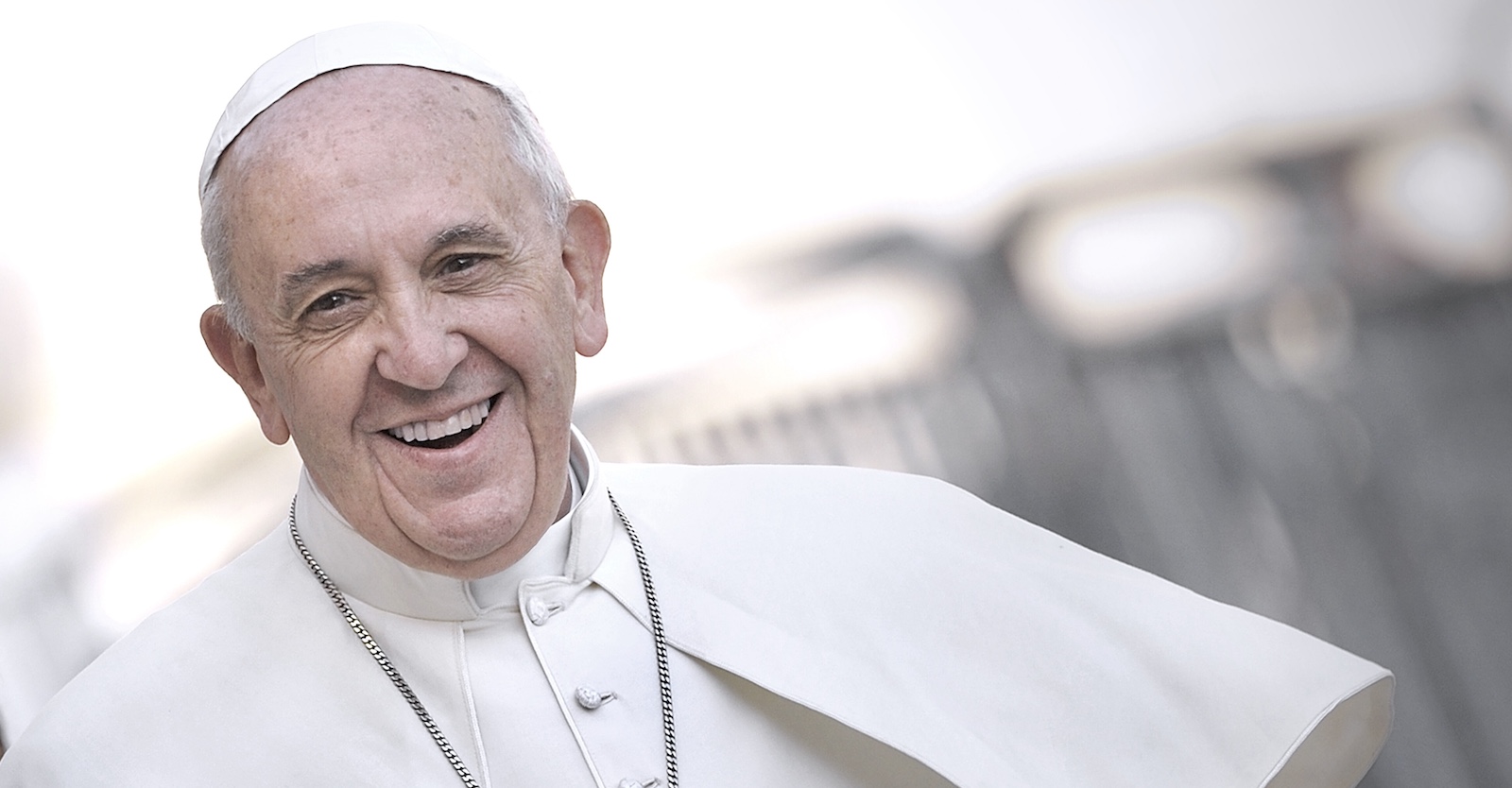 Porträt von Papst Franziskus aus dem Jahr 2013 mit freundlichem Lächeln im weißen Ornat im Freien, leicht seitlich aufgenommen.