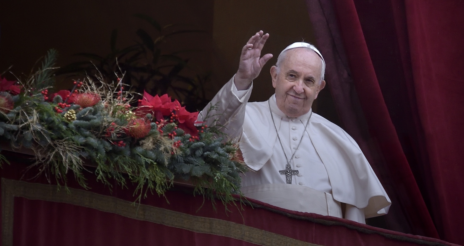 Papst Franziskus steht in weißem Gewand auf einem festlich geschmückten Balkon, winkt lächelnd in die Menge – im Hintergrund roter Vorhang und Tannenschmuck.