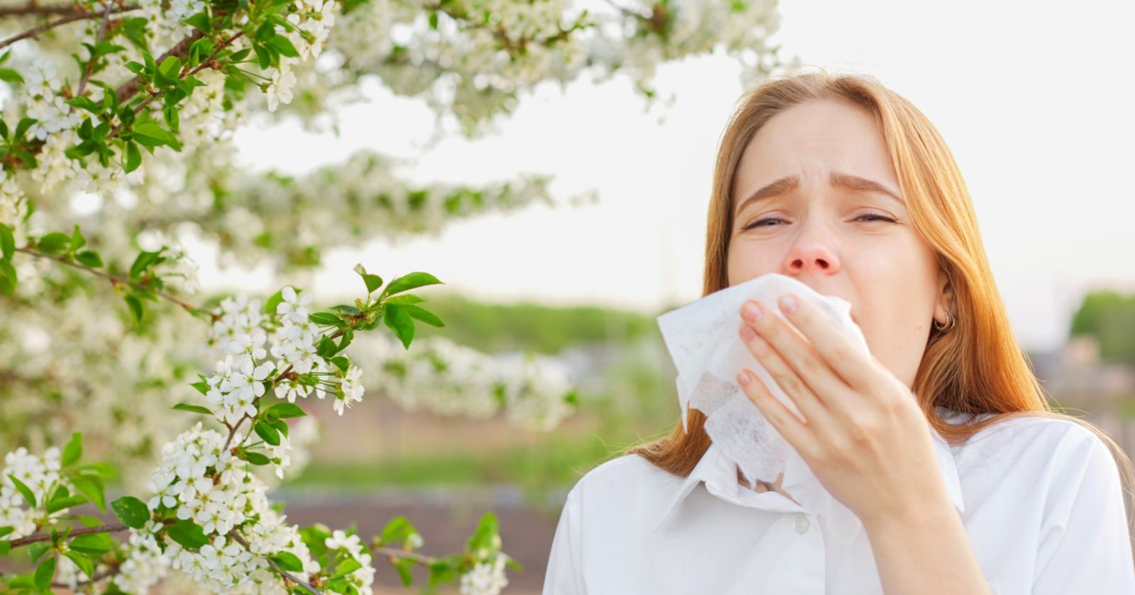 Rothaarige Frau niest im Freien in ein Taschentuch neben einem blühenden Strauch.