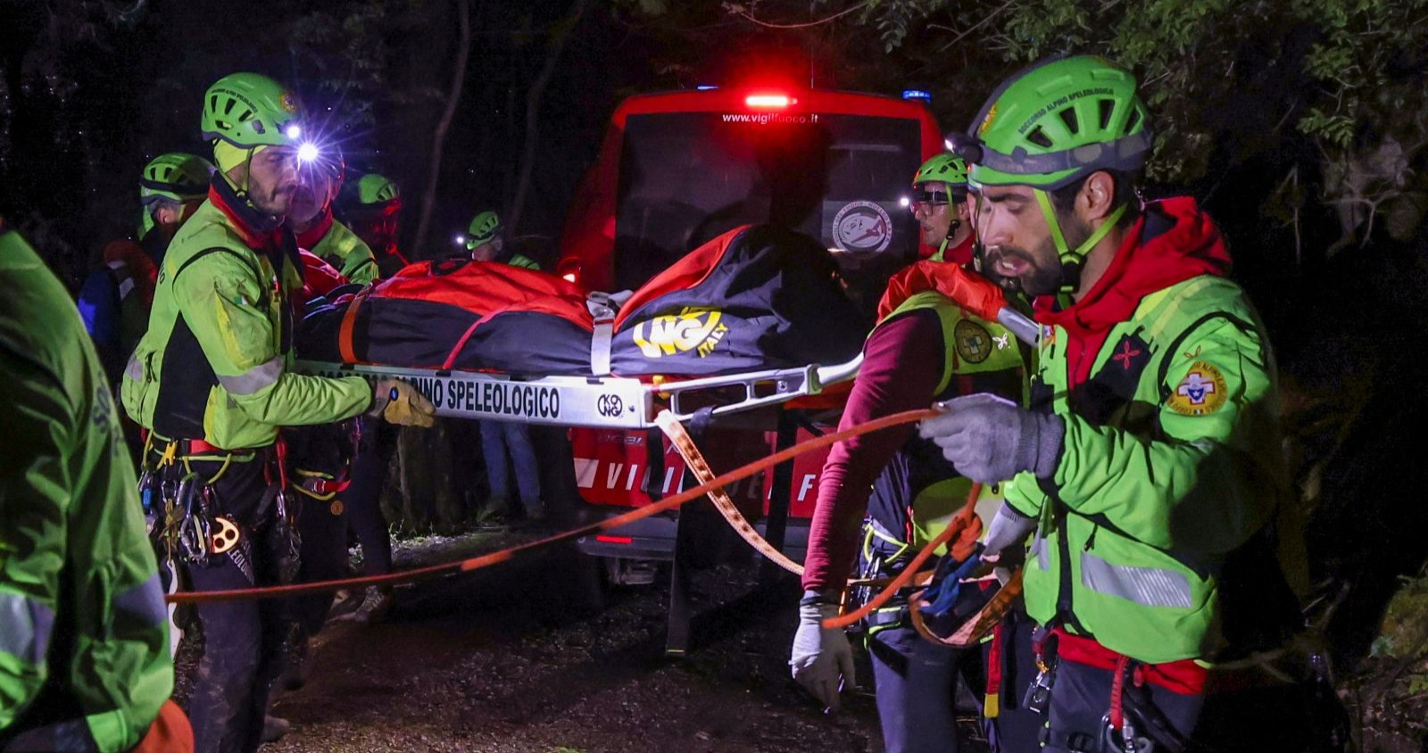 Rettungskräfte in grüner Schutzkleidung und Helmen transportieren bei Nacht eine Person auf einer Trage durch bewaldetes Gelände. Im Hintergrund steht ein Einsatzfahrzeug mit eingeschaltetem Blaulicht.