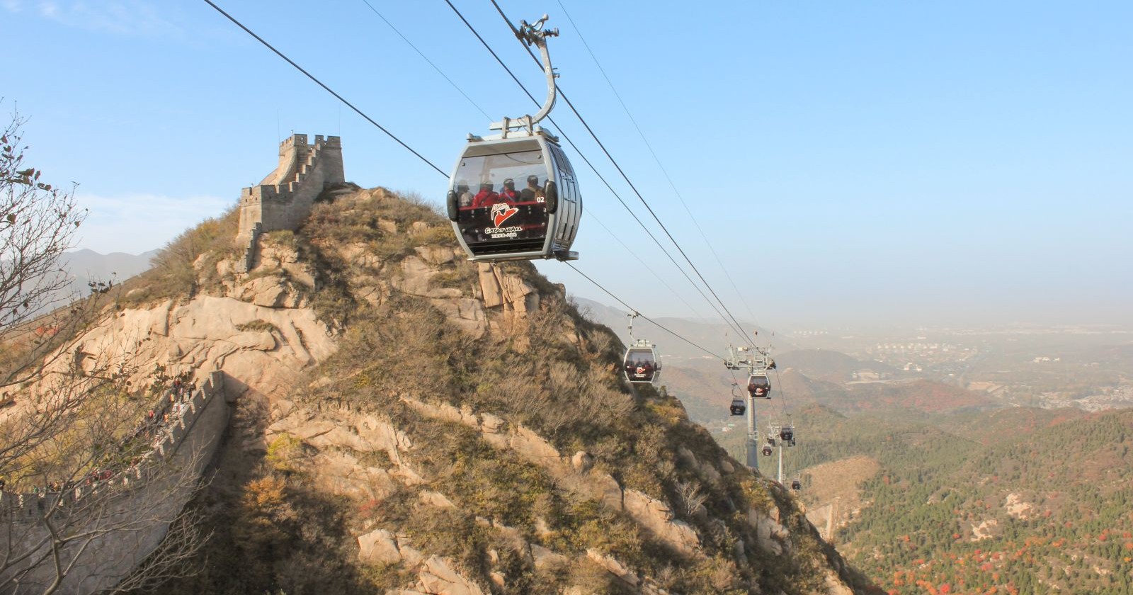 Eine Seilbahn schwebt vom Tal in die Höhe. Links im Bild ist die Chinesische Mauer zu sehen.
