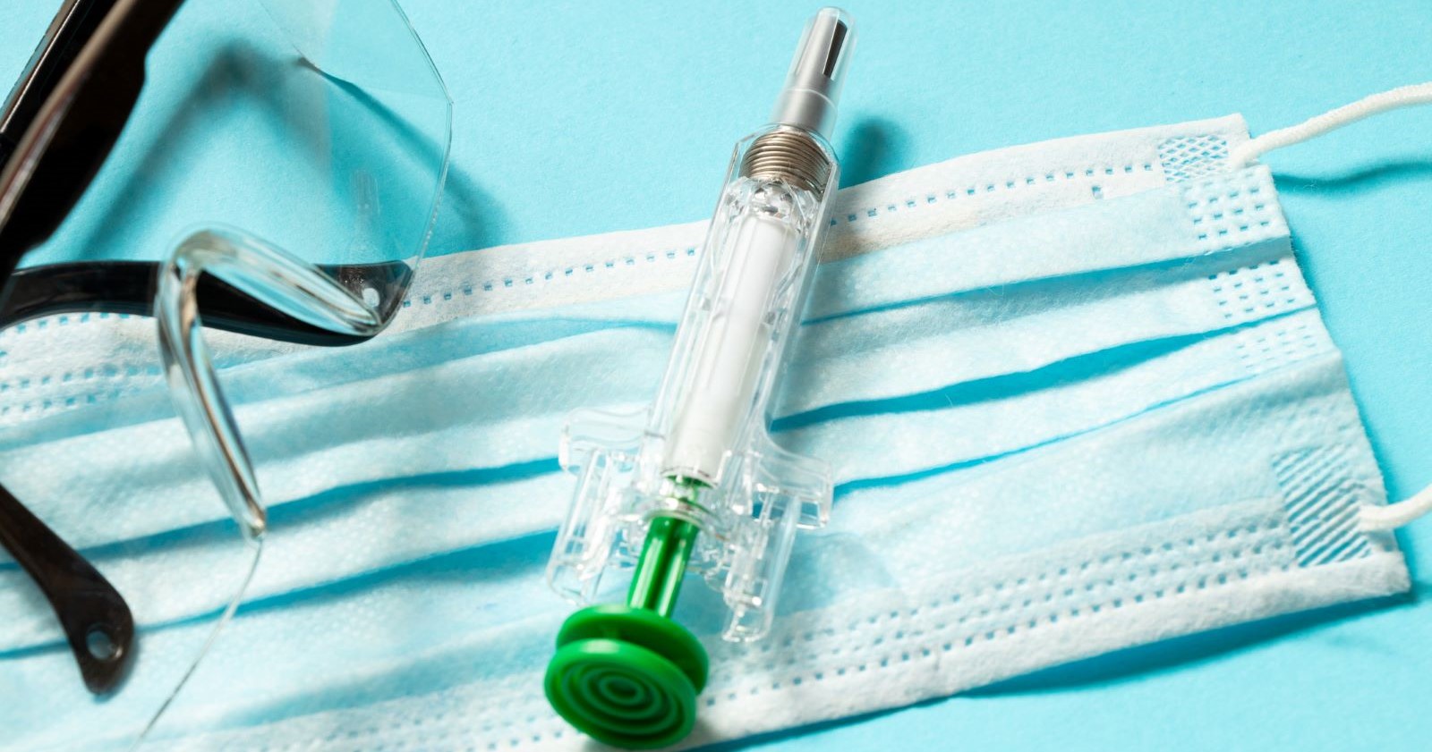 Medizinische Schutzmaske, Schutzbrille und transparente Spritze mit grünem Kolben auf hellblauem Hintergrund – Symbolbild für Pandemie, Impfschutz und Vorsorgemaßnahmen.