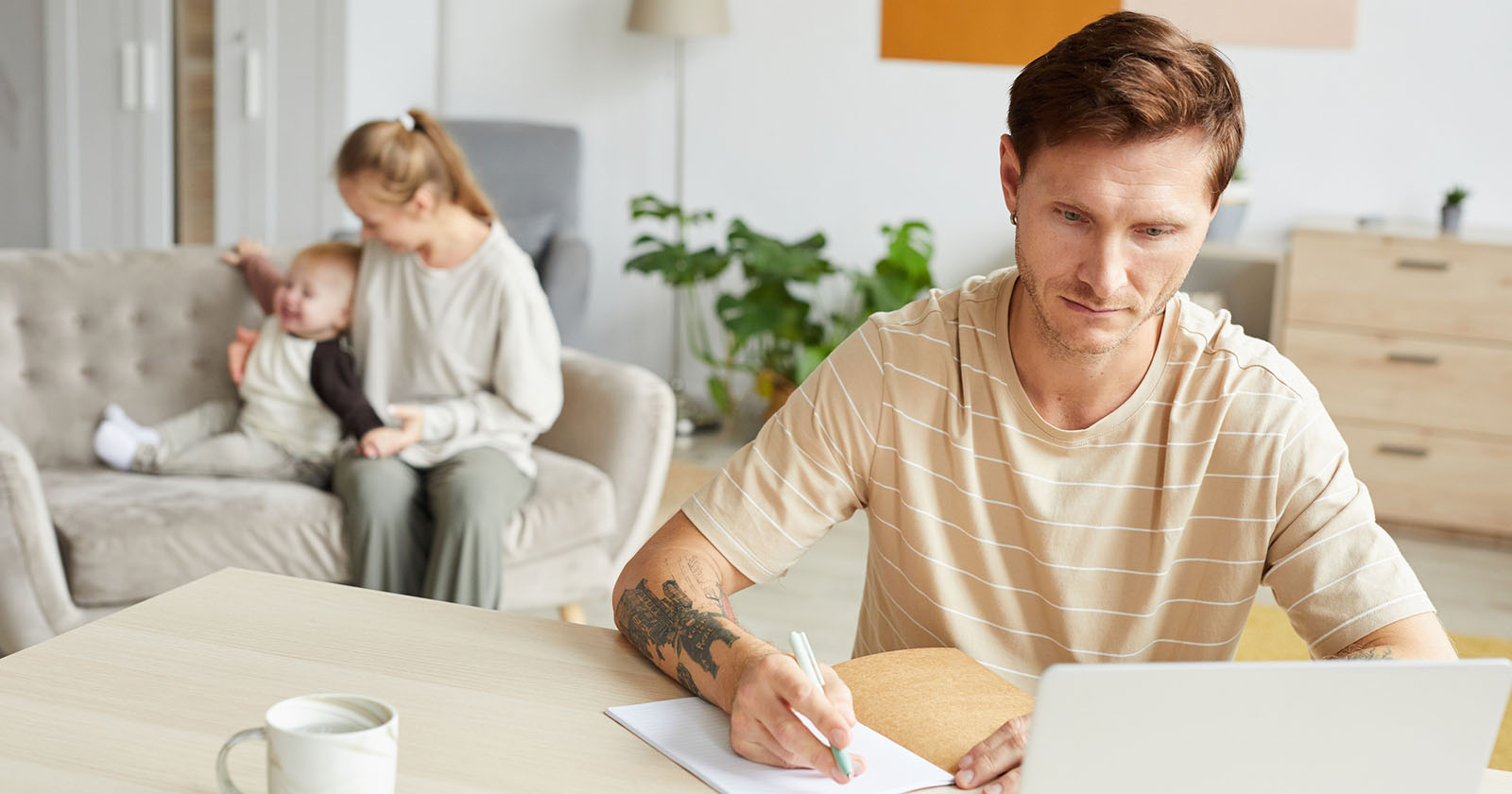 Junger Vater sitzt am Laptop am Küchentisch, während im Hintergrund seine Partnerin mit einem Kleinkind auf dem Sofa spielt